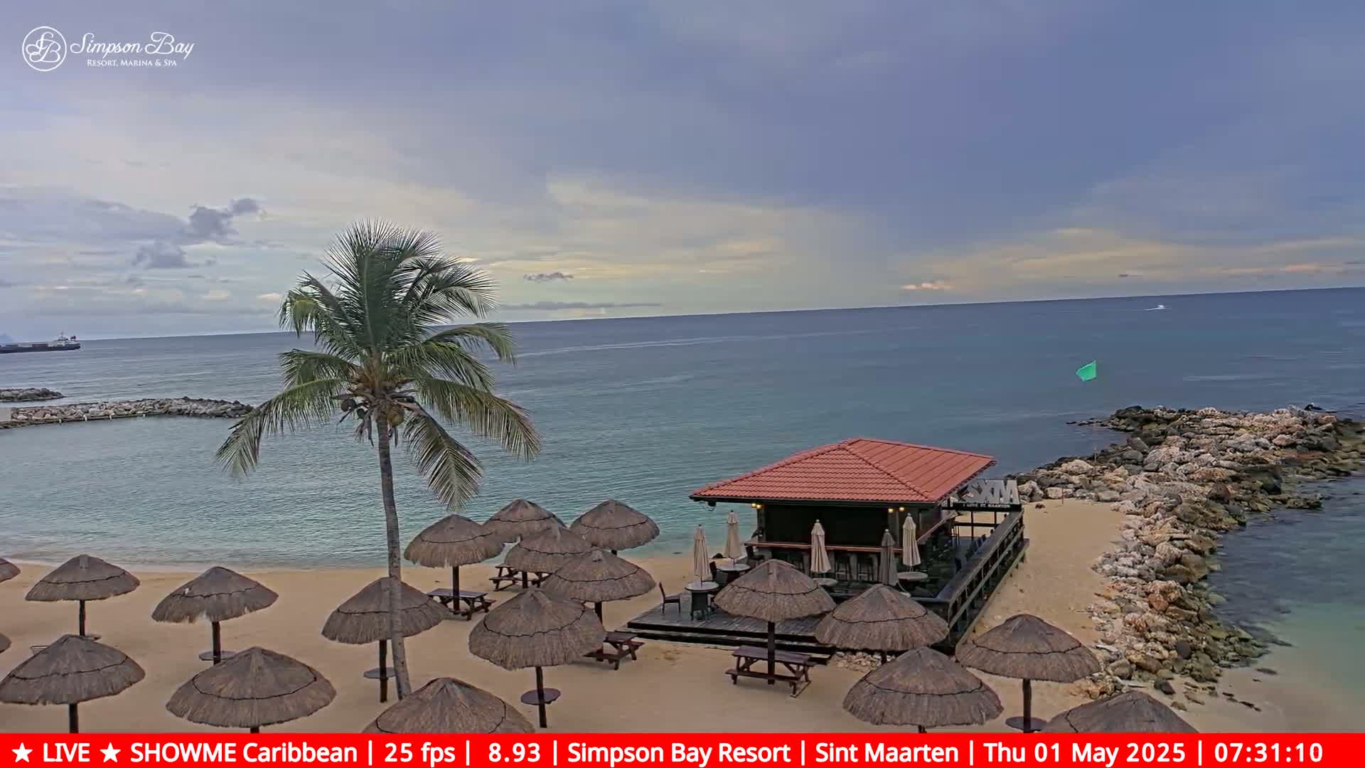 A sandy beach with thatched umbrellas, picnic tables, and a small bar overlooks a calm ocean under a mostly cloudy sky.