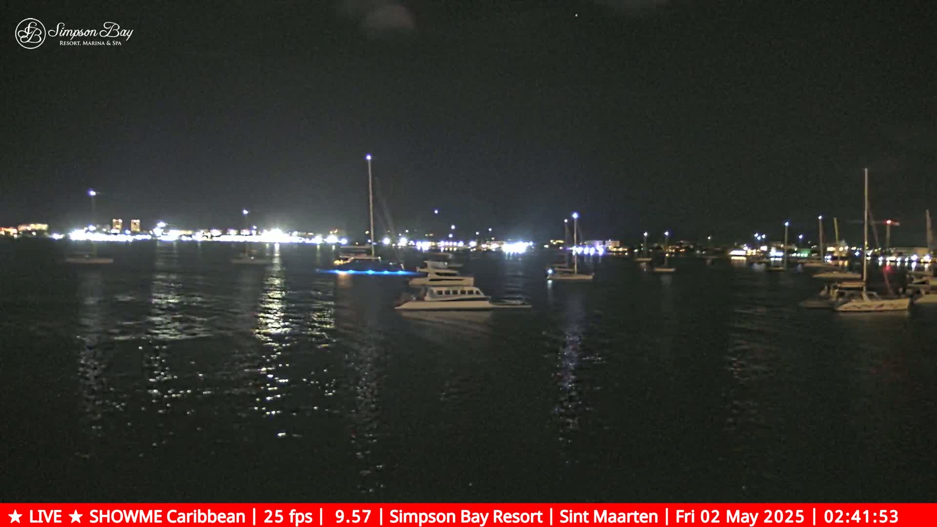 At night, numerous boats are moored in a calm body of water, illuminated by lights from the waterfront buildings and the boats themselves.