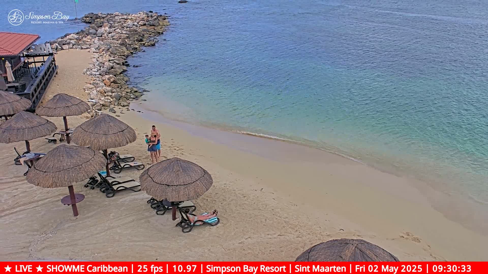 A sandy beach with calm, turquoise water, thatched umbrellas, lounge chairs, and a few people, is viewed from above.