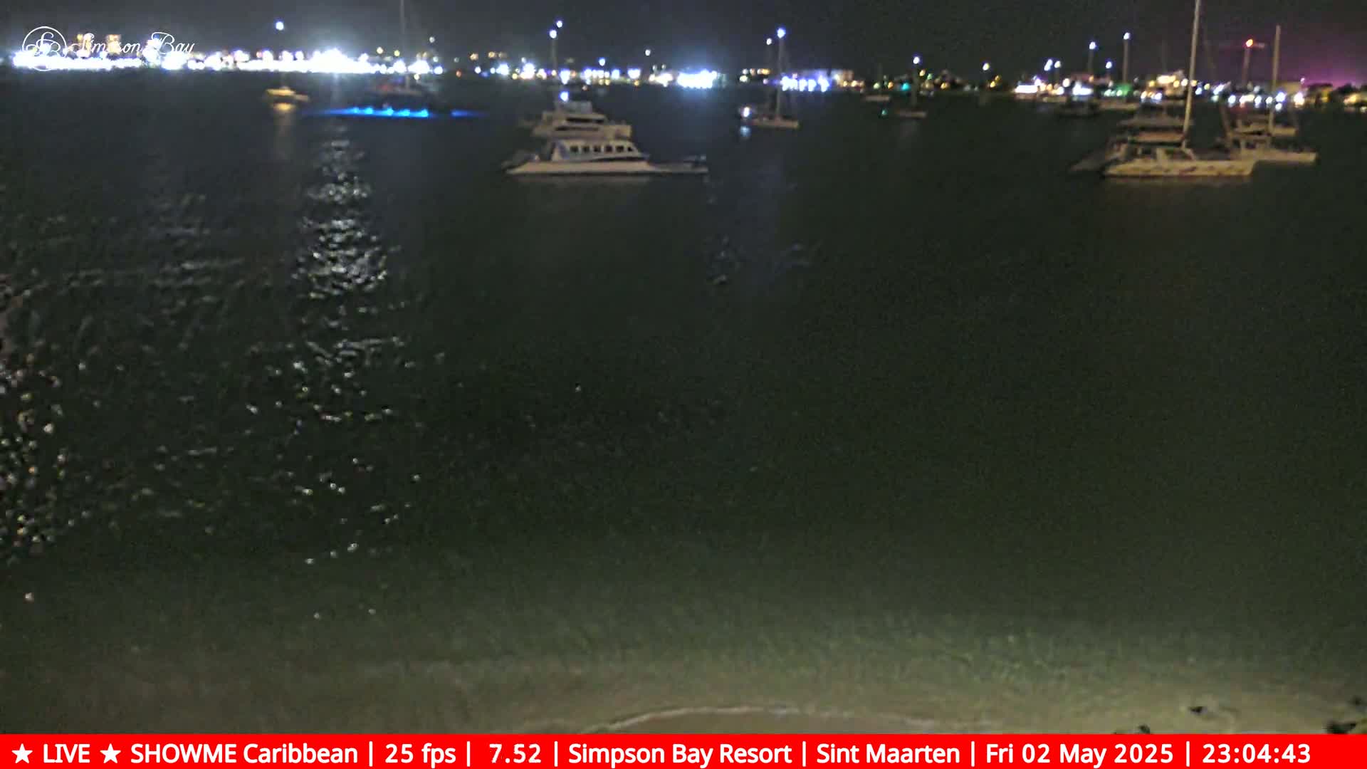Several boats are moored in a dark, calm body of water at night, illuminated by lights from a nearby shore.