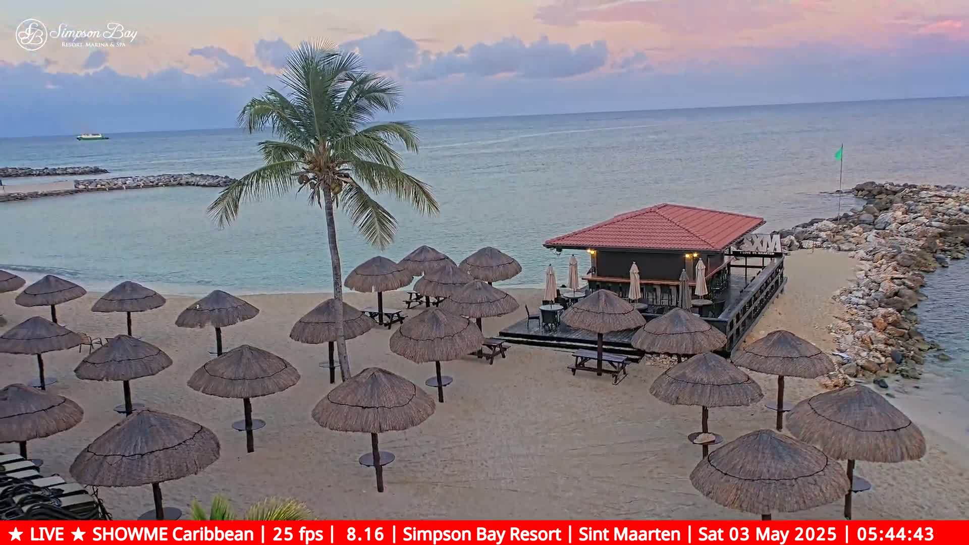 A beach bar with thatched umbrellas sits on a sandy beach next to calm ocean water under a partly cloudy, twilight sky.