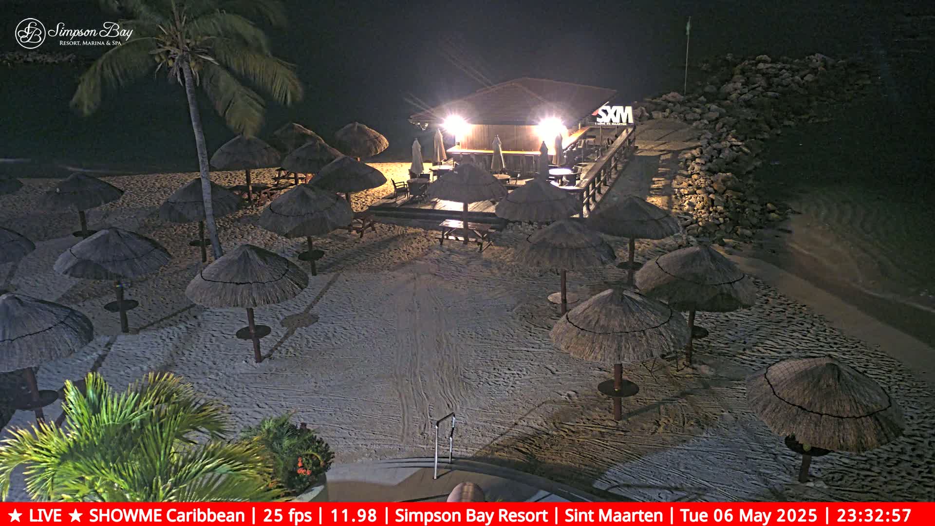 A nighttime beach scene shows numerous thatched umbrellas on a sandy beach, illuminated by the lights of a beachfront bar.