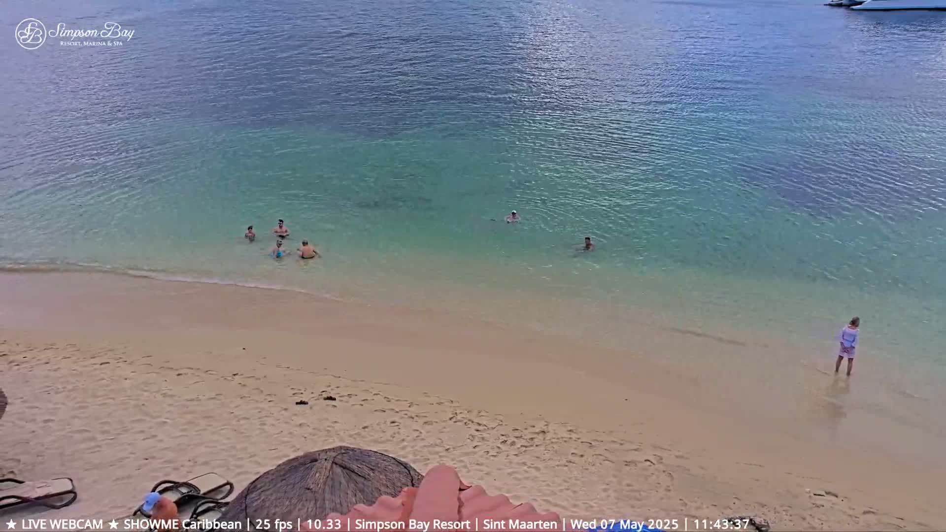 A sandy beach slopes gently into calm, shallow turquoise water where several people are swimming and wading.