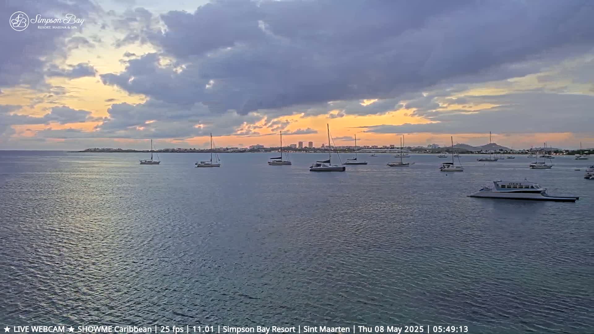 Numerous sailboats and a ferry are moored in calm water at sunset under a partly cloudy sky.