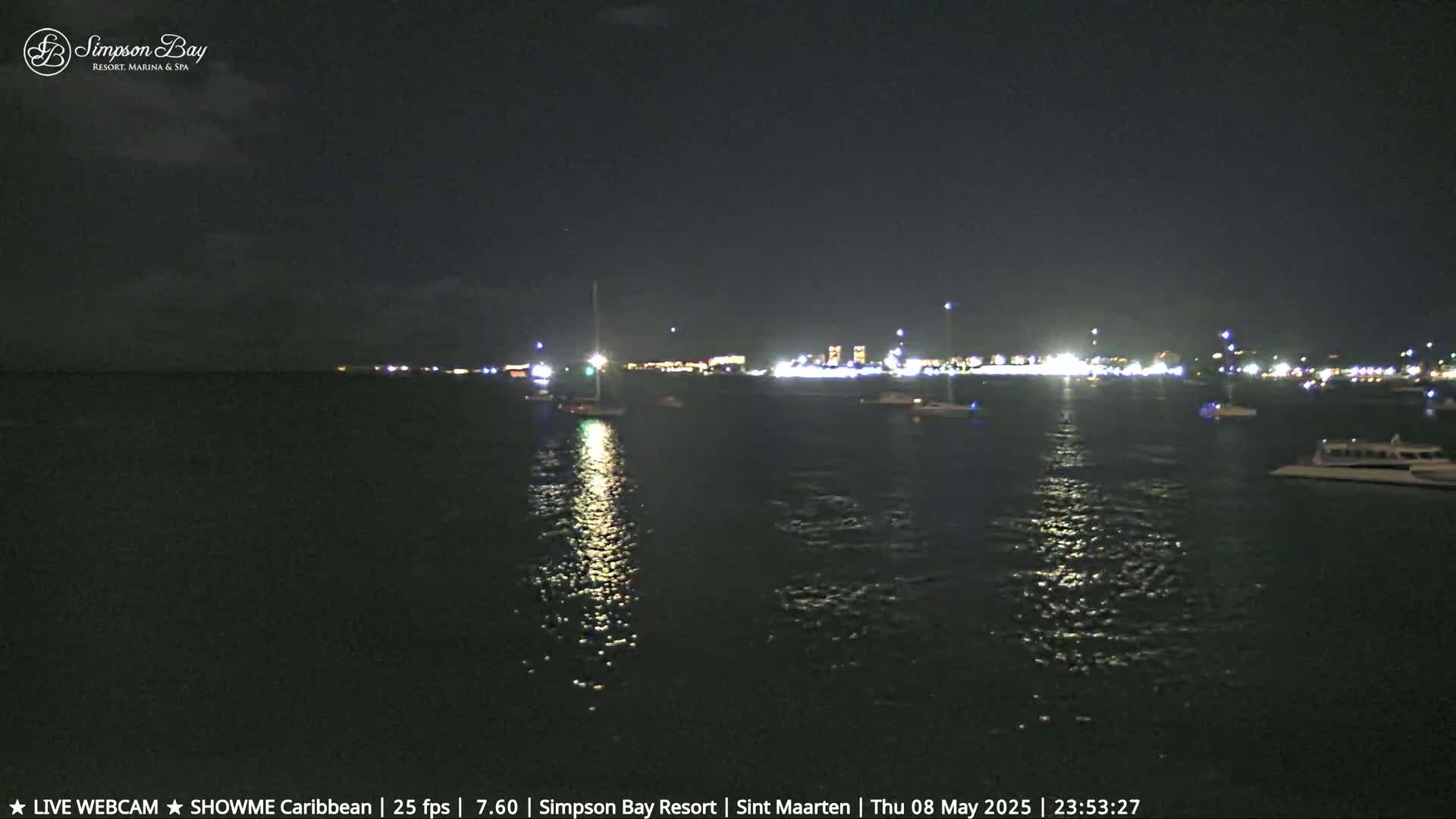 At night, a calm body of water reflects the lights from a distant shoreline where boats are moored.