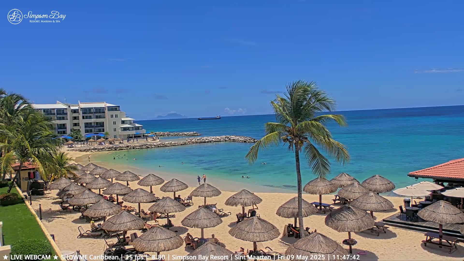 A sandy beach with numerous thatched umbrellas, a resort building, and calm turquoise water under a clear blue sky.