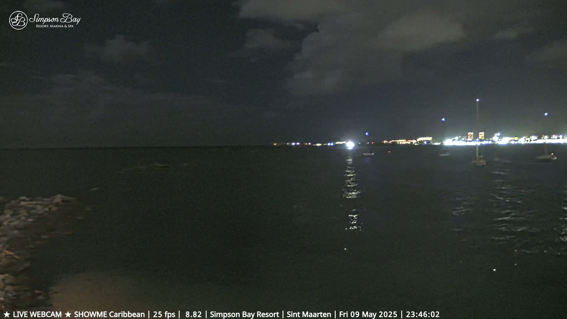 A nighttime view of a calm body of water with a few boats and a brightly lit shoreline under a partly cloudy sky.