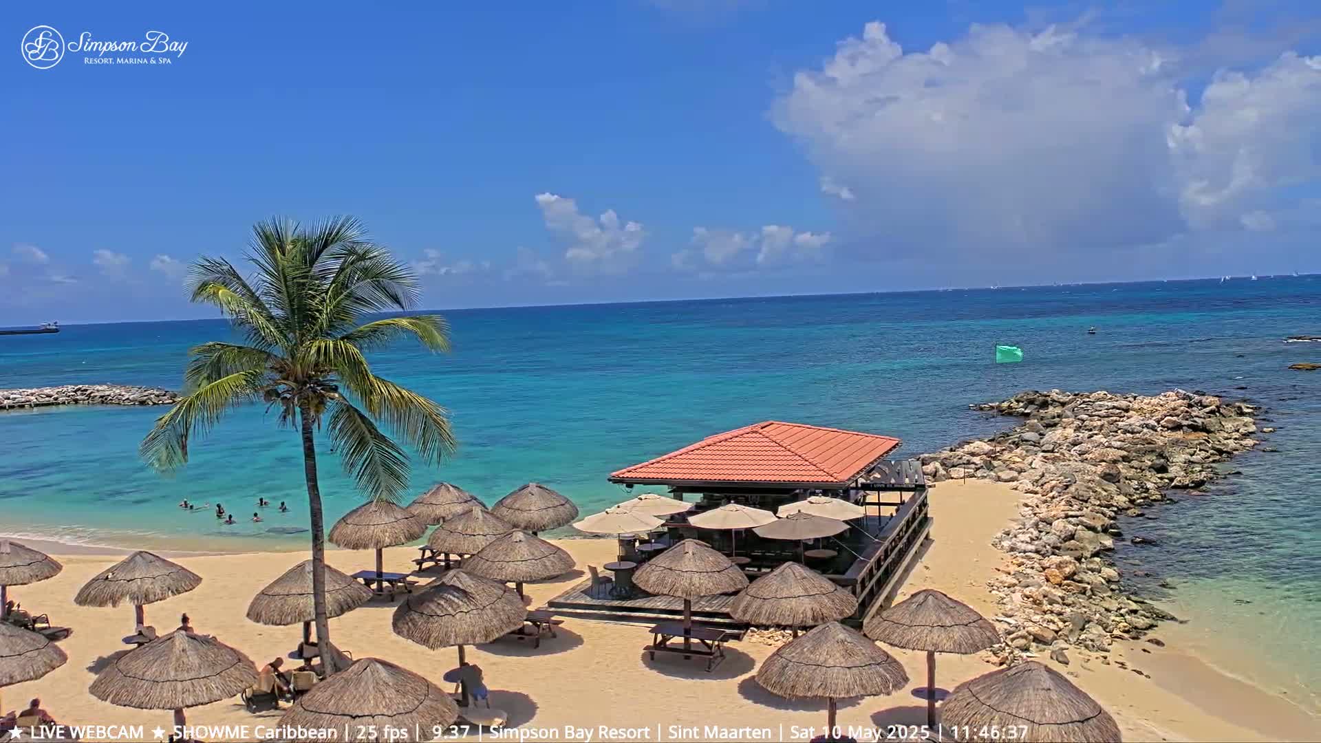 A sandy beach with thatched umbrellas, a beachfront bar, and a palm tree, under a partly cloudy blue sky, overlooks a calm, turquoise ocean.