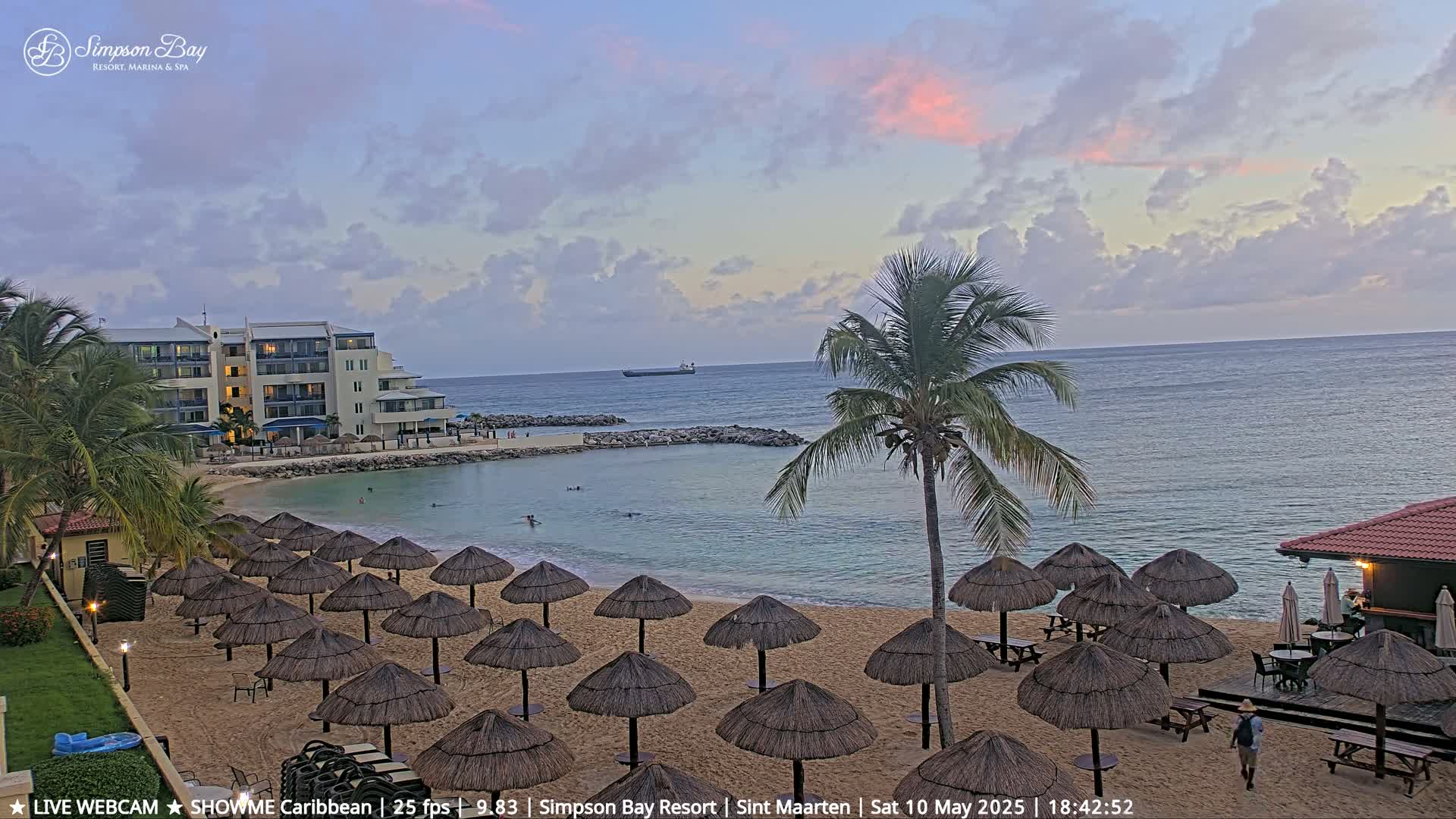 A sandy beach with numerous thatched umbrellas, a calm bay, a resort building, and a palm tree is visible under a partly cloudy sunset sky.