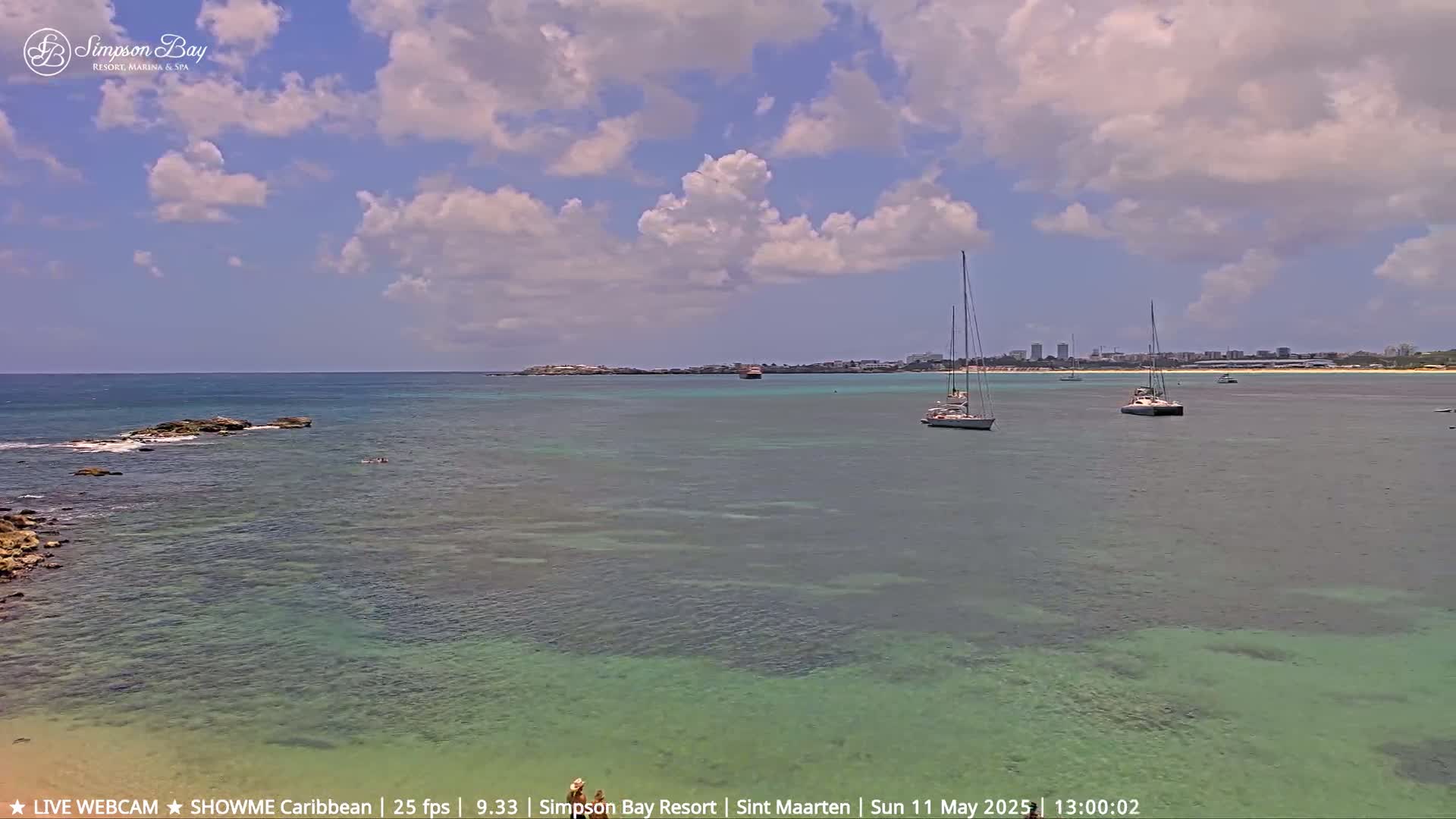 A partly cloudy day shows a tranquil bay with several sailboats anchored, shallow clear water, and a distant shoreline with buildings under a mostly sunny sky.