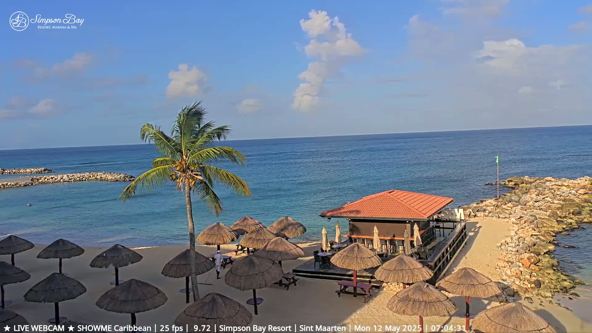 A sandy beach with thatched umbrellas, a small beach bar, and a palm tree under a partly cloudy blue sky next to a calm ocean.