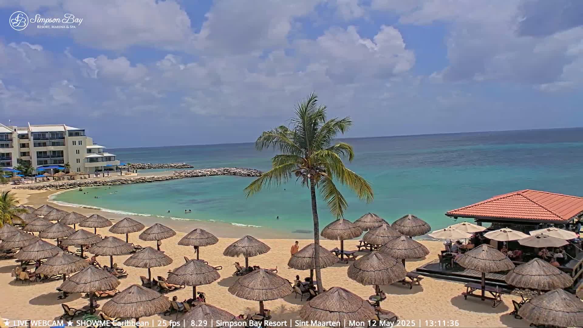 A sandy beach with numerous thatched umbrellas, a beachfront bar, and a calm, turquoise ocean under a partly cloudy sky.