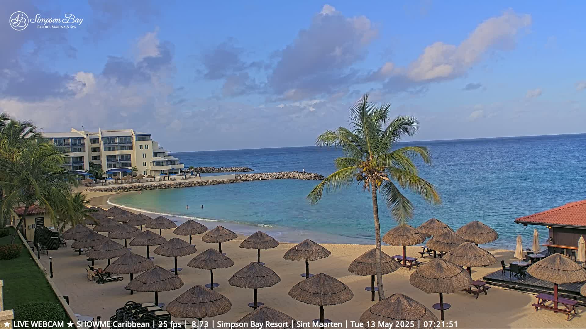 A sandy beach with numerous thatched umbrellas, a resort building, and a calm ocean under a partly cloudy sky.