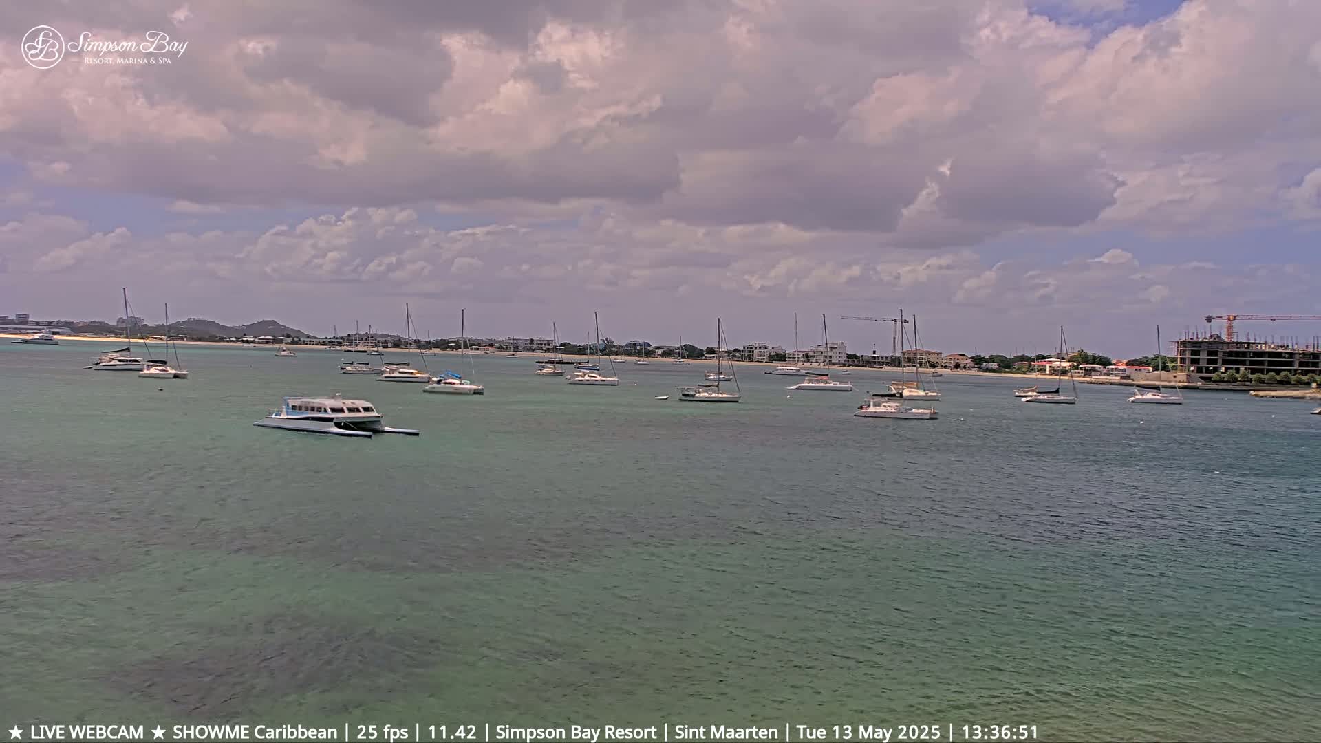 Several sailboats and motorboats are anchored in a calm, shallow bay under a partly cloudy sky.