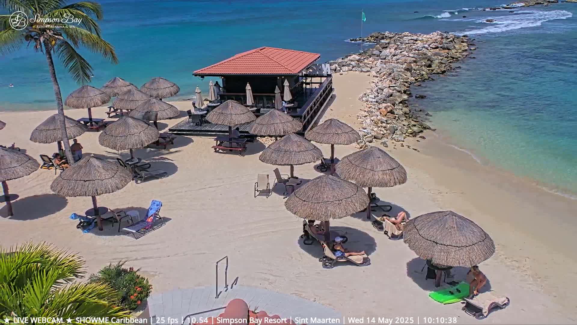 A beach bar sits on a sandy beach with numerous thatched umbrellas and lounge chairs under a sunny sky, next to a calm, teal ocean.