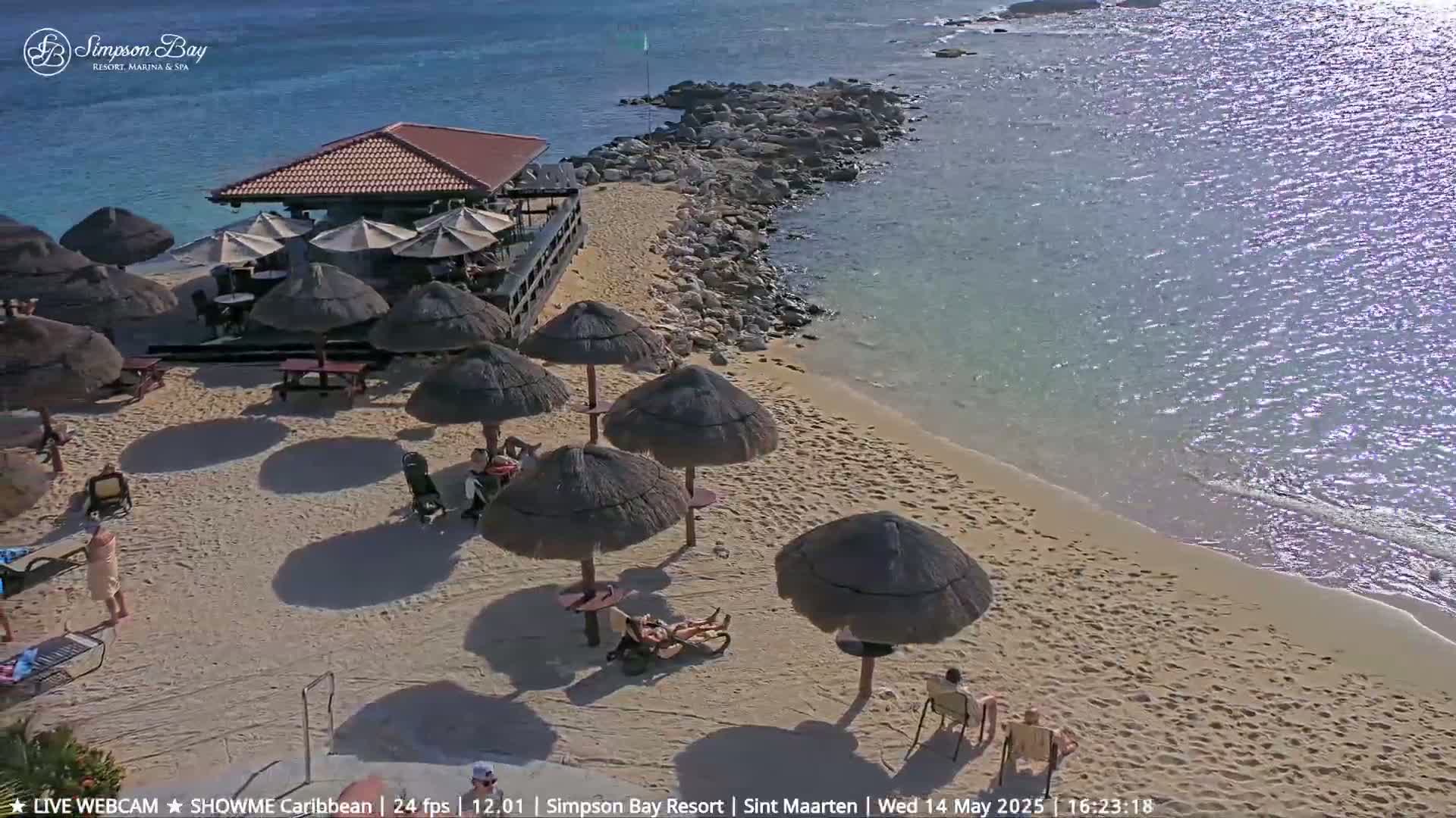 A sandy beach with thatched umbrellas and beachgoers is adjacent to a calm, clear ocean and a small beachfront building.