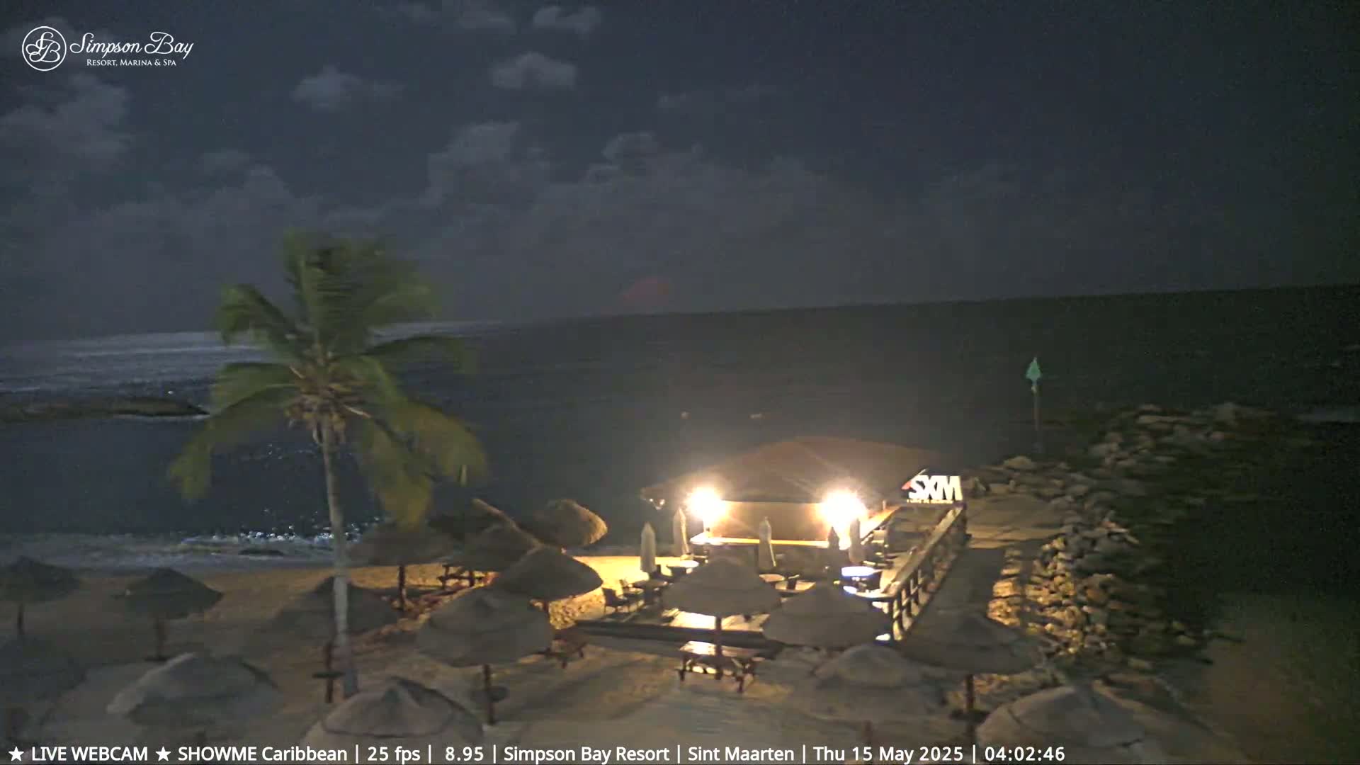 A nighttime beach scene shows a lit beachfront bar and several beach umbrellas under a partly cloudy sky.