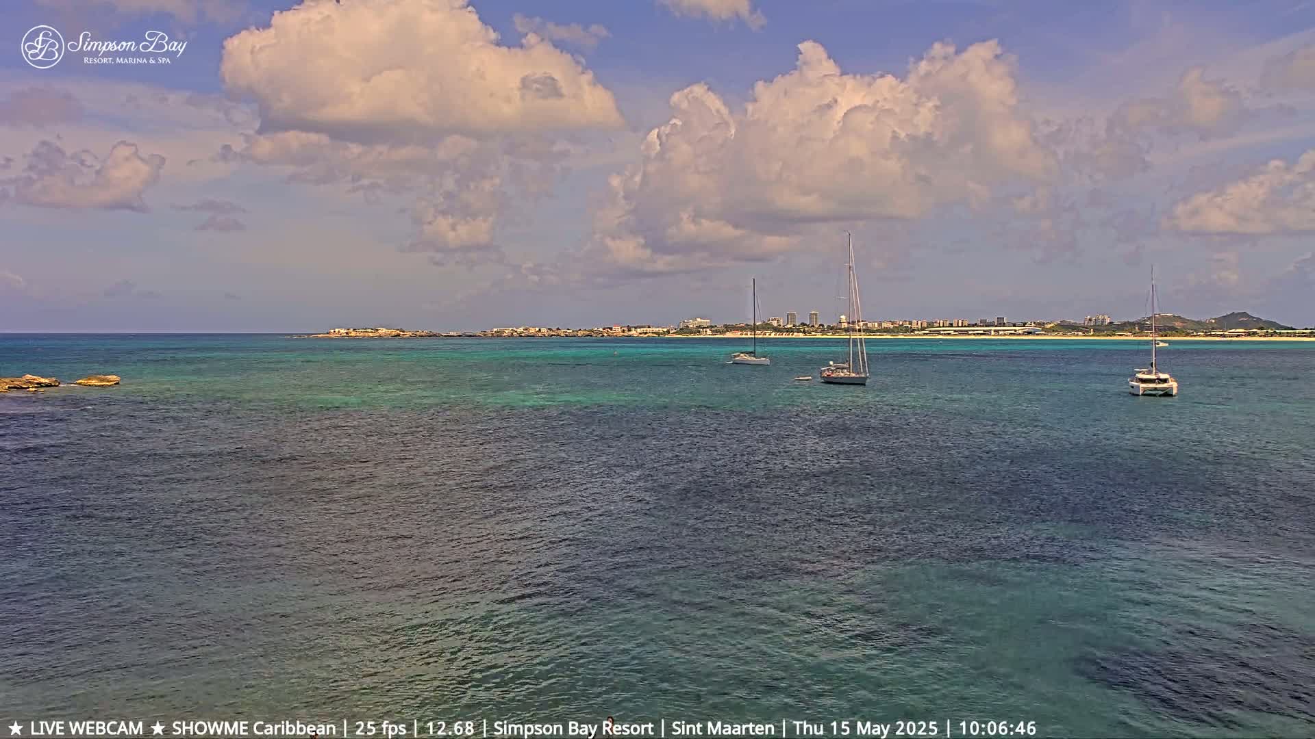 A partly cloudy day shows a tranquil bay with three sailboats anchored near a distant coastline under a mostly sunny sky.