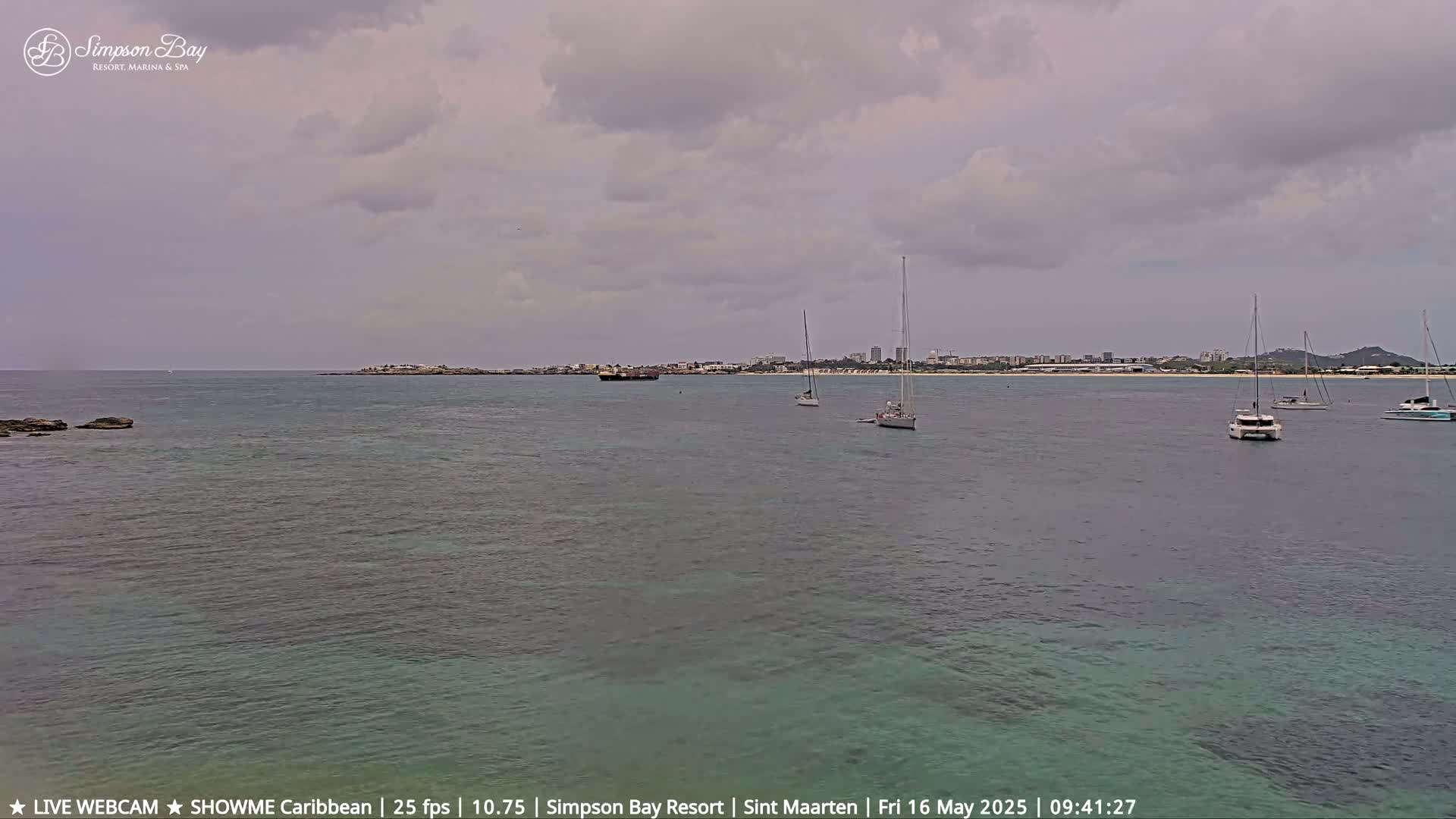 A calm bay with several sailboats and a distant shoreline under a cloudy sky.