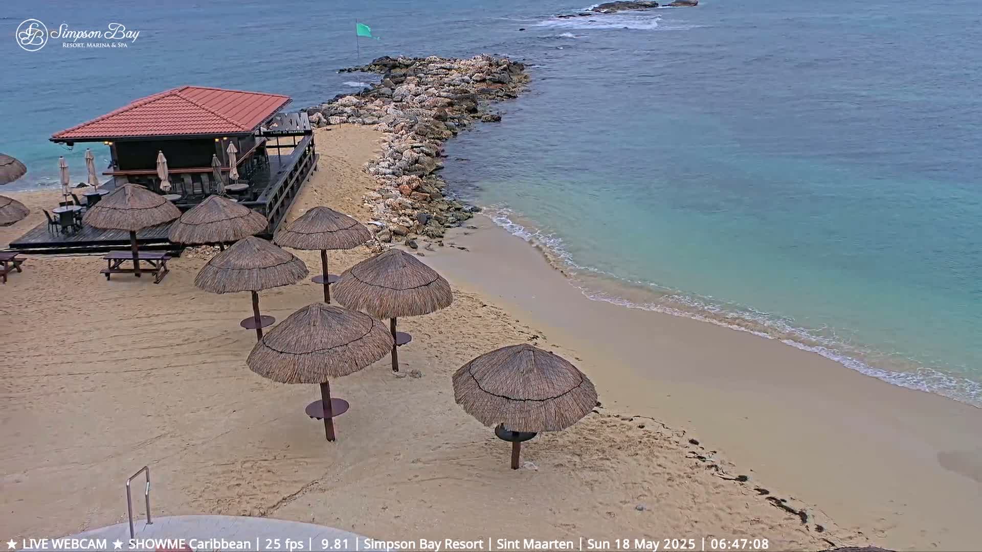 A beach bar with thatched umbrellas sits on a sandy beach next to calm, turquoise water and a rocky outcropping.