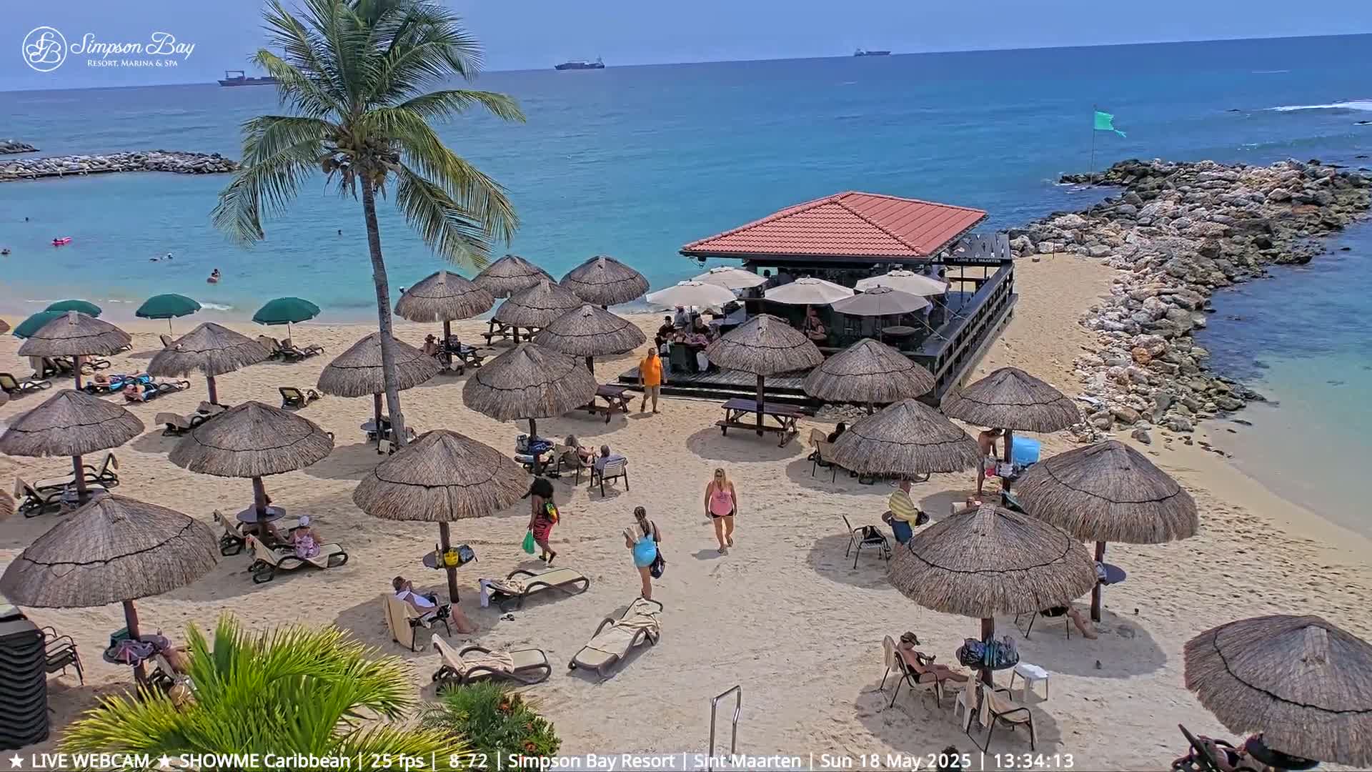 A sunny beach scene shows numerous thatched umbrellas shading lounge chairs and people on a sandy beach next to a small beachfront bar and calm turquoise water.