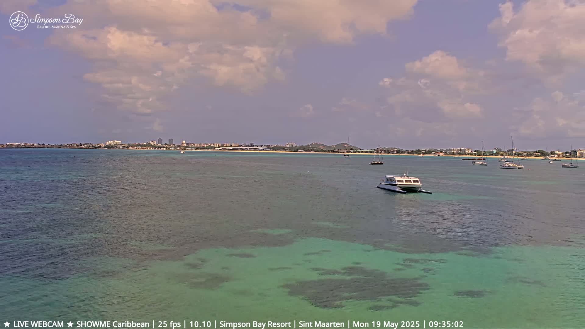 A partly cloudy day reveals a calm, turquoise bay with several boats and a distant shoreline featuring buildings and hills.