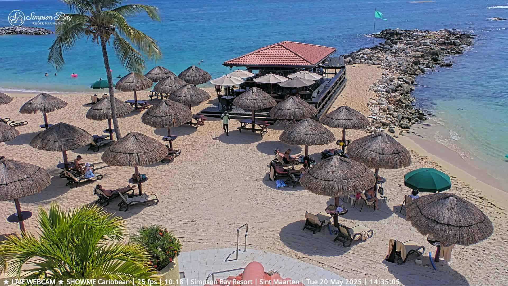 A sandy beach with numerous thatched umbrellas, lounge chairs, and a beachfront bar, all under a sunny sky, next to a calm, clear ocean.