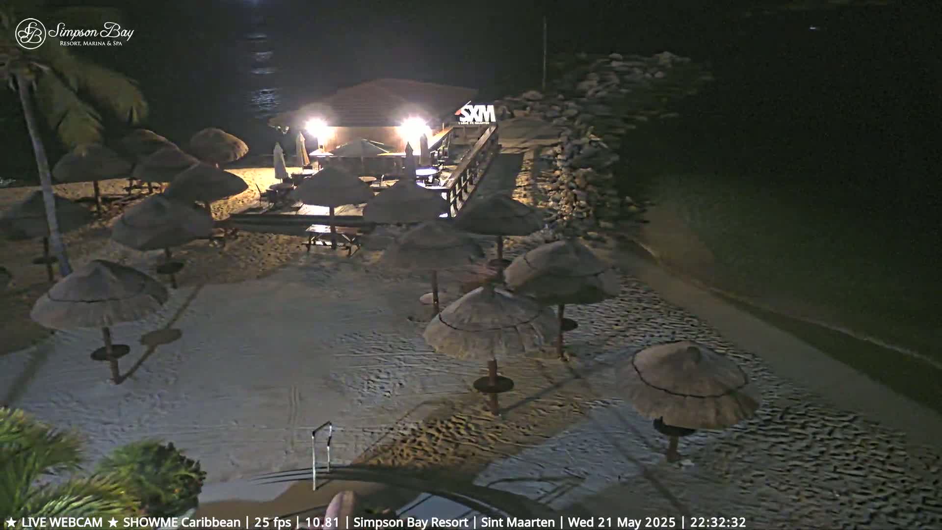 A nighttime beach scene shows numerous thatched umbrellas on a sandy beach near a lit beachfront building and calm water.