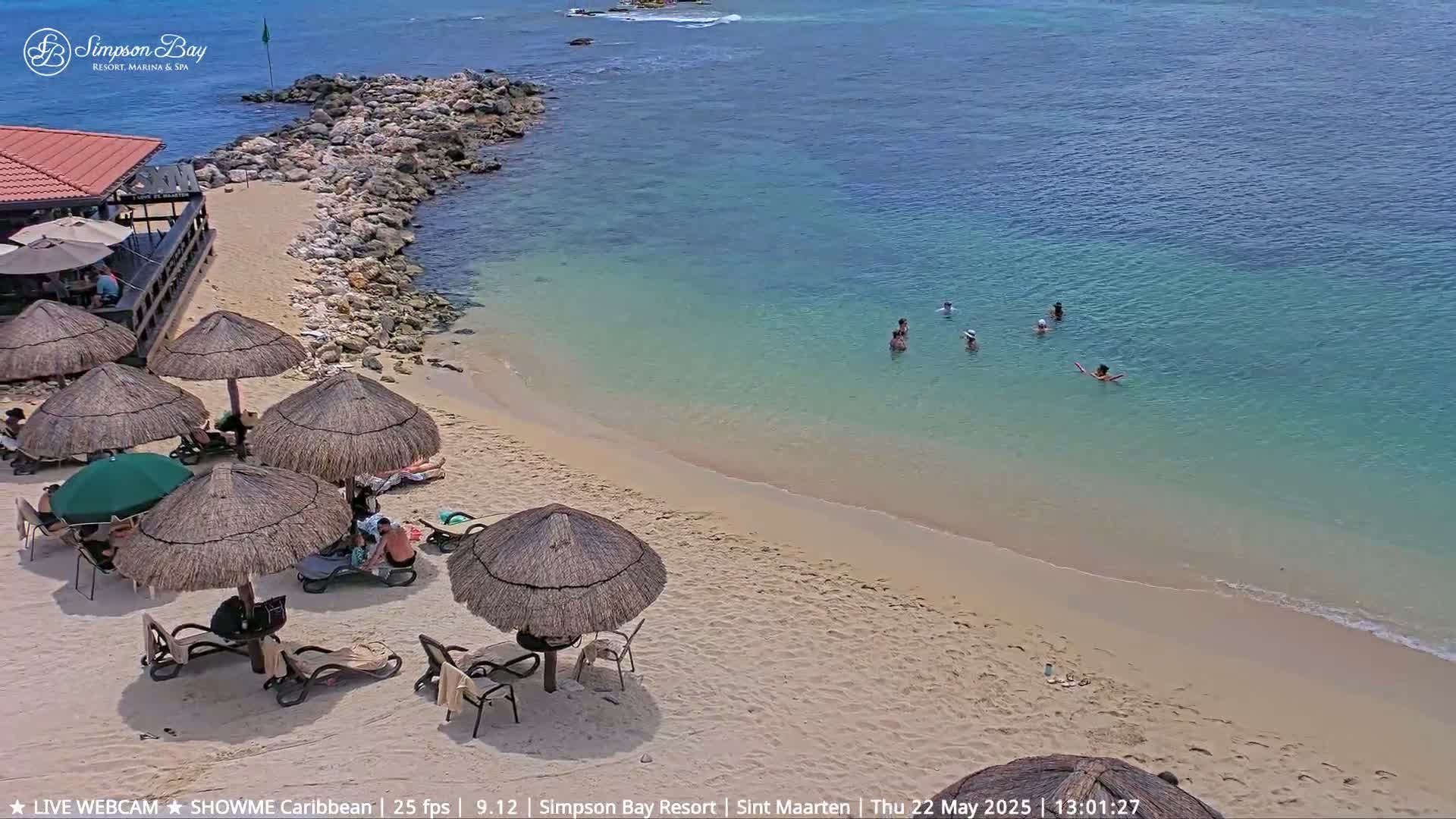 A sandy beach with thatched umbrellas and lounge chairs, several people swimming in clear turquoise water, under a sunny sky.
