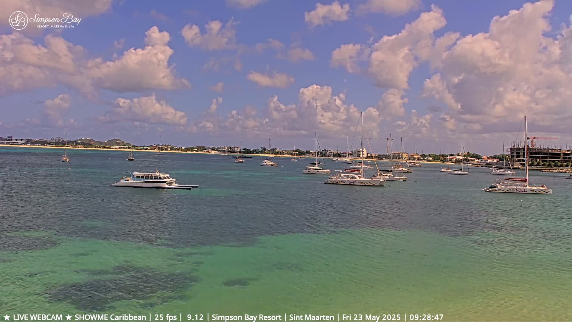 A partly cloudy day shows a calm bay with numerous sailboats and a ferry docked near a shoreline with buildings and construction.