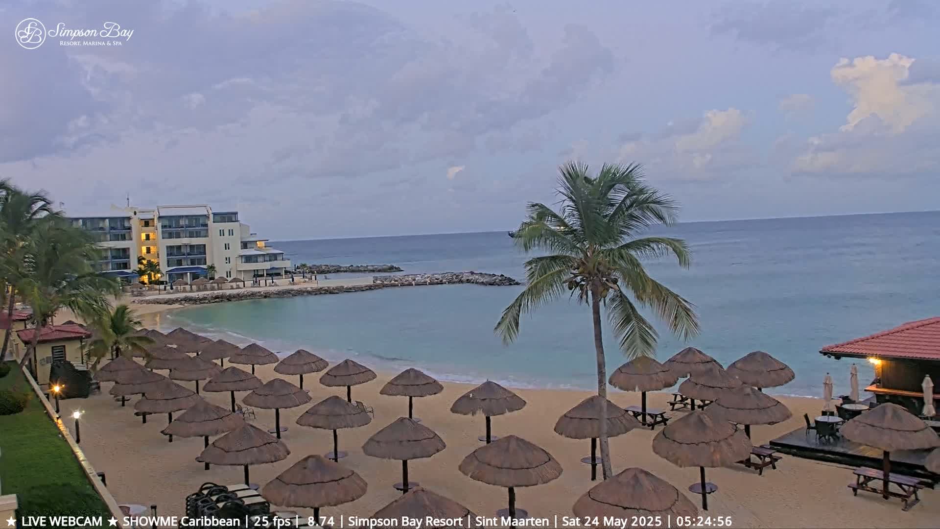 A sandy beach with numerous thatched umbrellas and patio furniture is situated in front of a resort building under a partly cloudy sky.