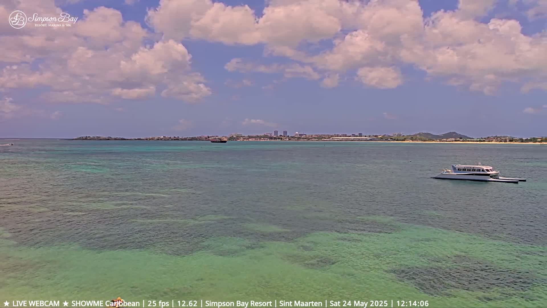A partly cloudy day shows a calm, shallow, turquoise body of water with a white catamaran in the distance, a landmass with buildings visible in the far background.