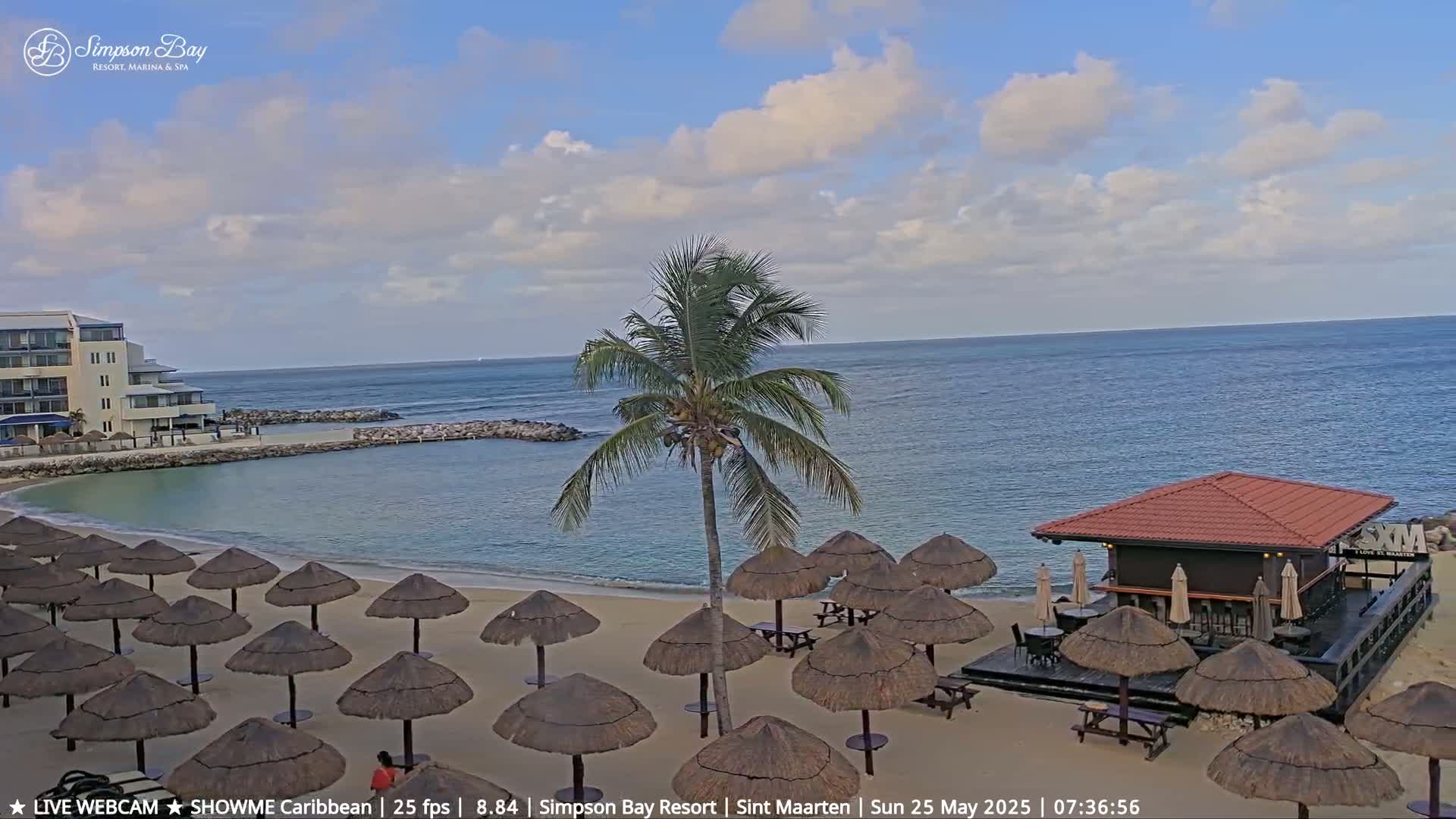 A partly cloudy day at a beach resort shows a sandy beach with numerous thatched umbrellas, a beachfront bar, a calm ocean, and a resort building in the background.