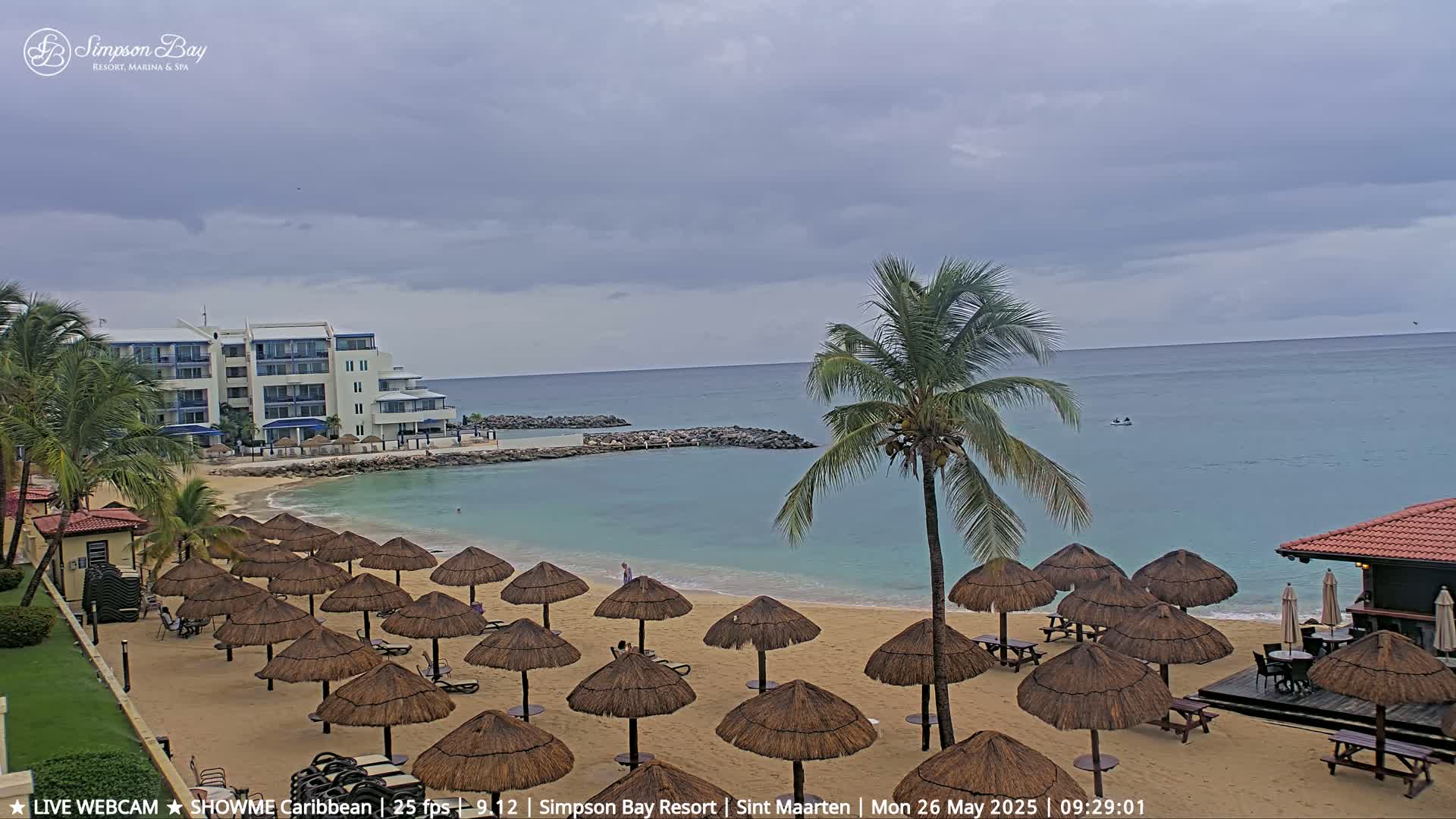 A sandy beach with numerous thatched umbrellas, a calm turquoise ocean, and a resort building under an overcast sky.