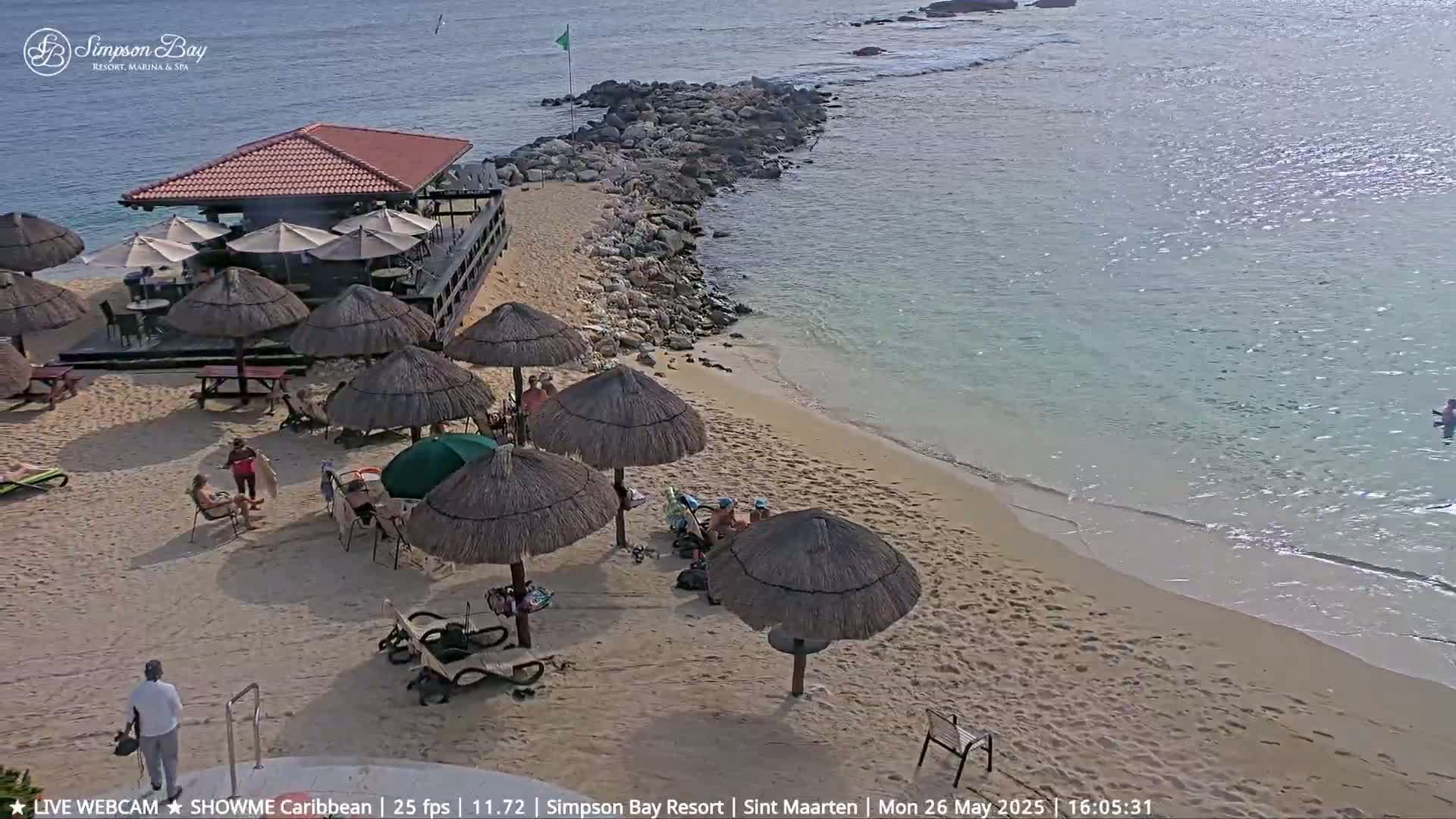 A sandy beach with thatched umbrellas, beach chairs, and a beachfront bar is shown on a sunny day, with calm, clear water visible beyond a rocky shoreline.