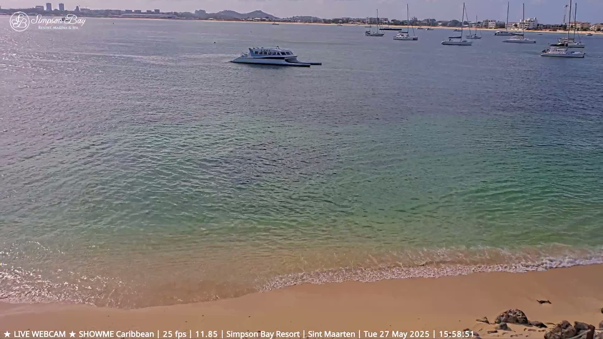 A sandy beach meets calm, shallow turquoise water with several boats anchored in the distance under a sunny sky.