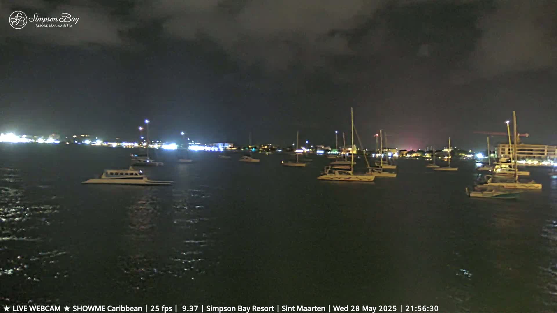 At night, under a dark, cloudy sky, numerous boats are moored in a calm body of water near a brightly lit shoreline.