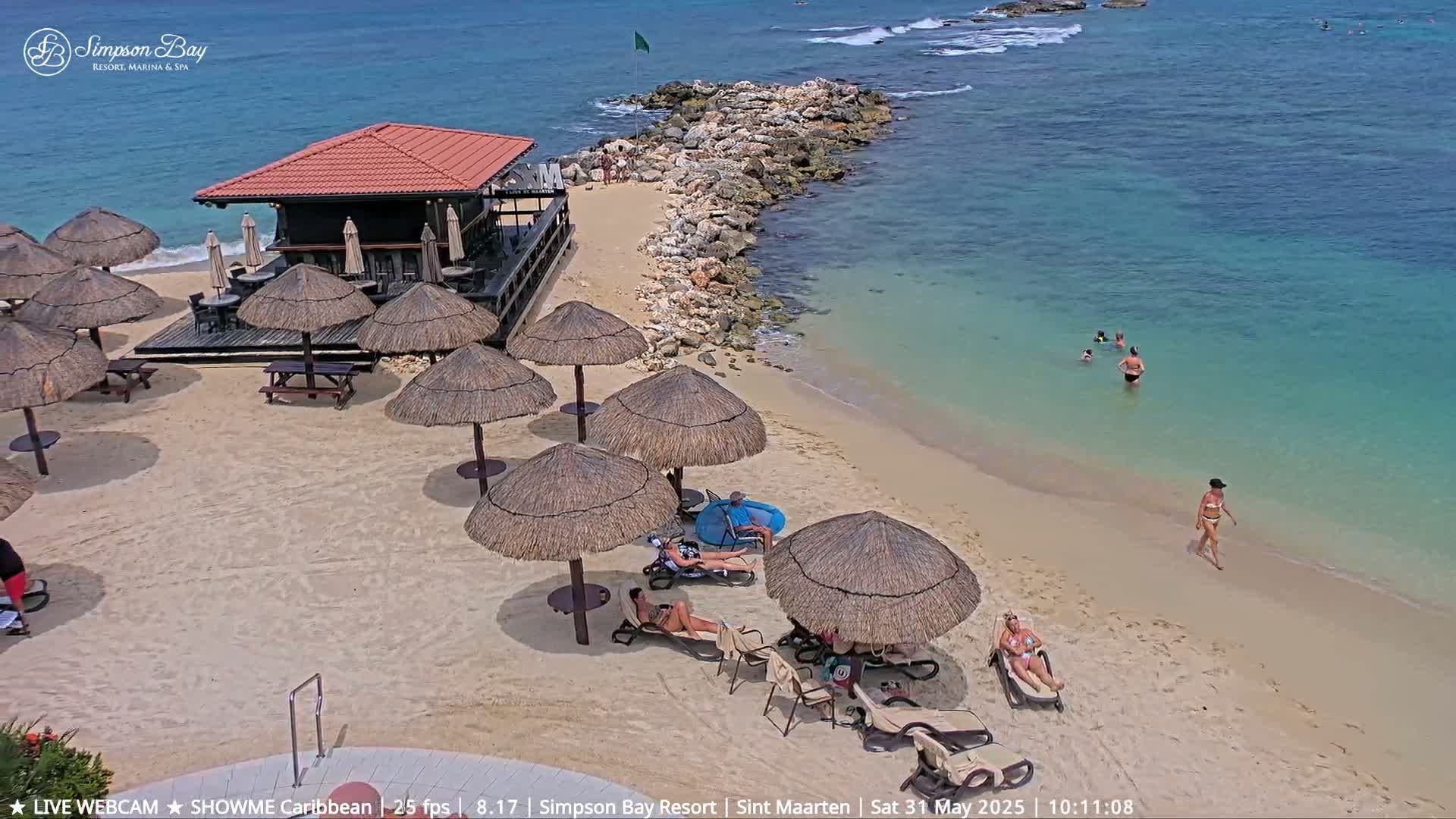 A beach bar and numerous thatched umbrellas on a sandy beach next to a rocky point, with people swimming and sunbathing in clear, shallow turquoise water under a sunny sky.