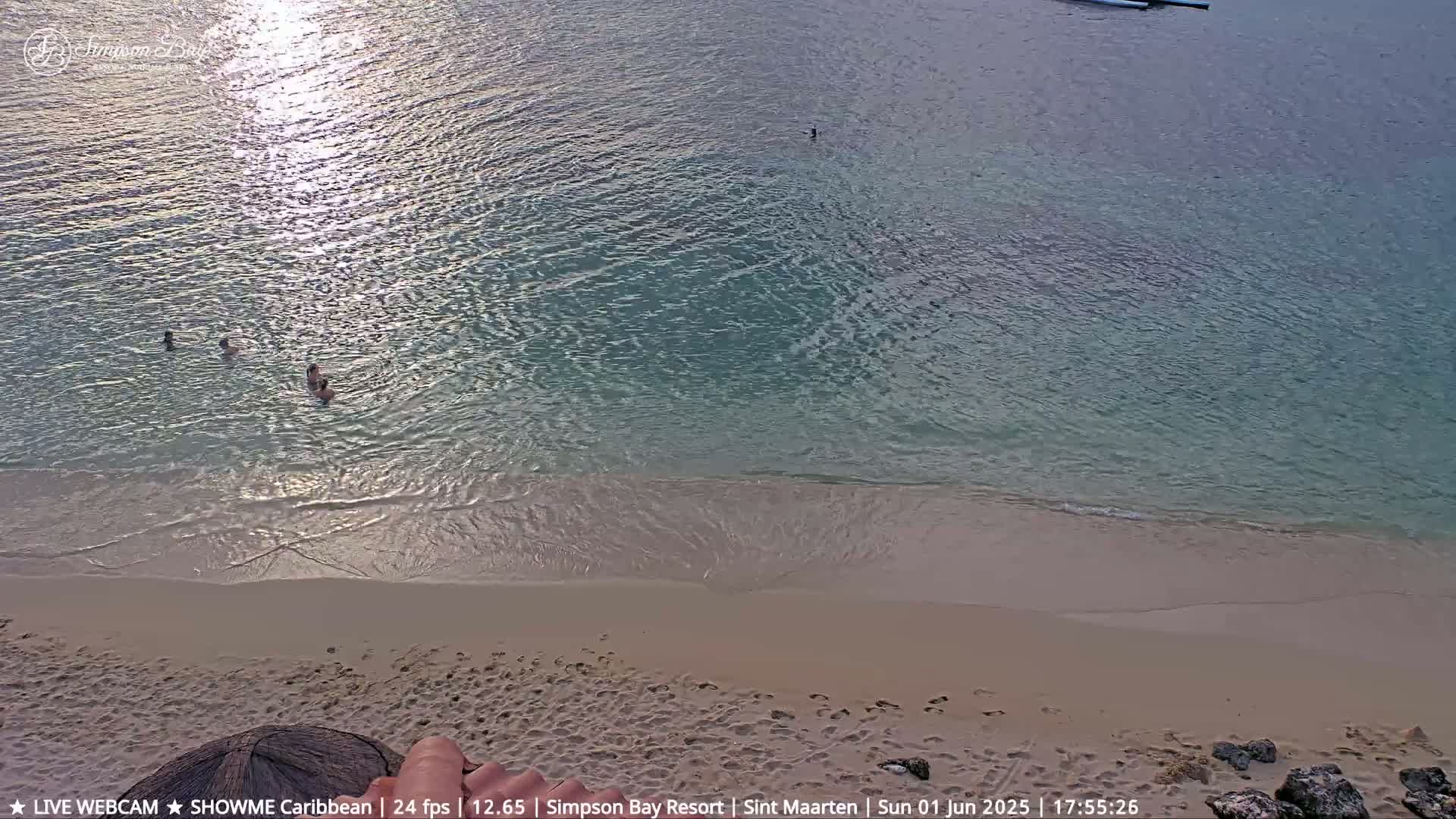 Sunlight glints on calm, shallow turquoise water lapping a sandy beach where several people are swimming.