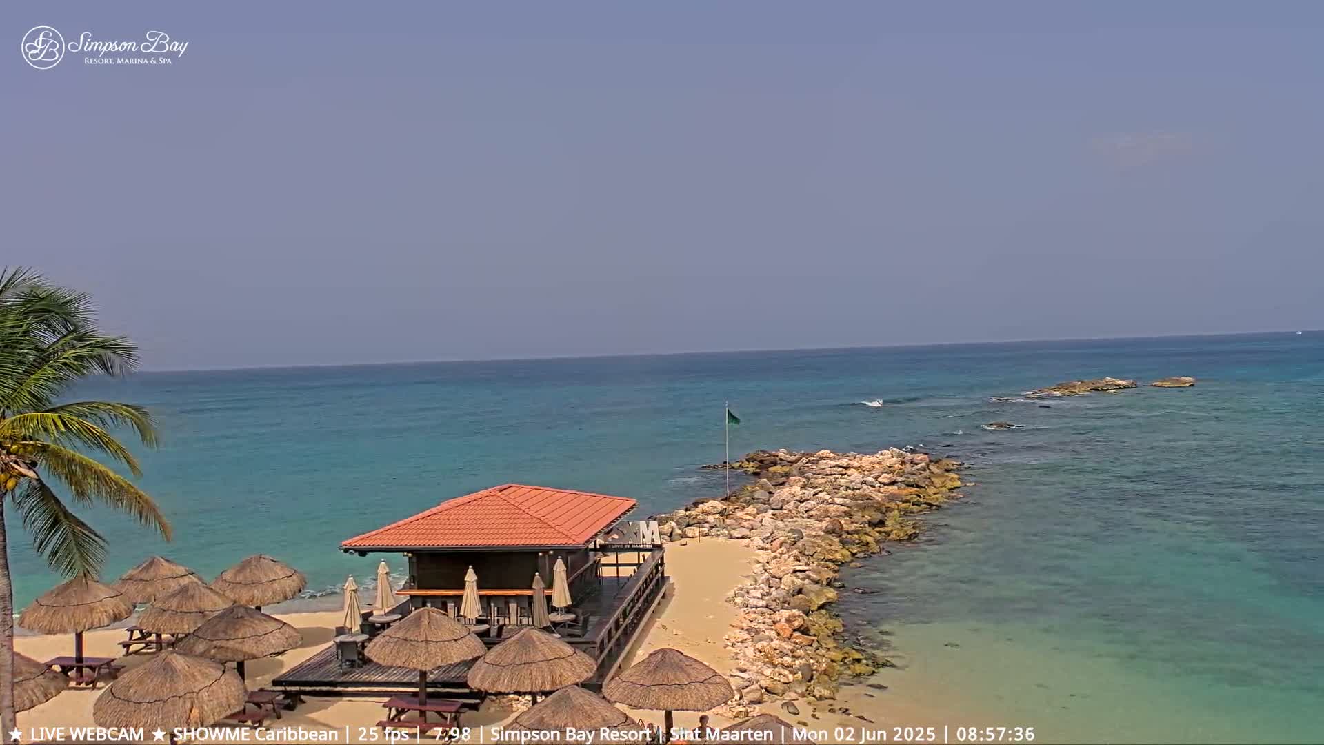 A small, red-roofed beach bar sits on a sandy shore next to a rocky outcrop, with turquoise water extending to the horizon under a clear sky.