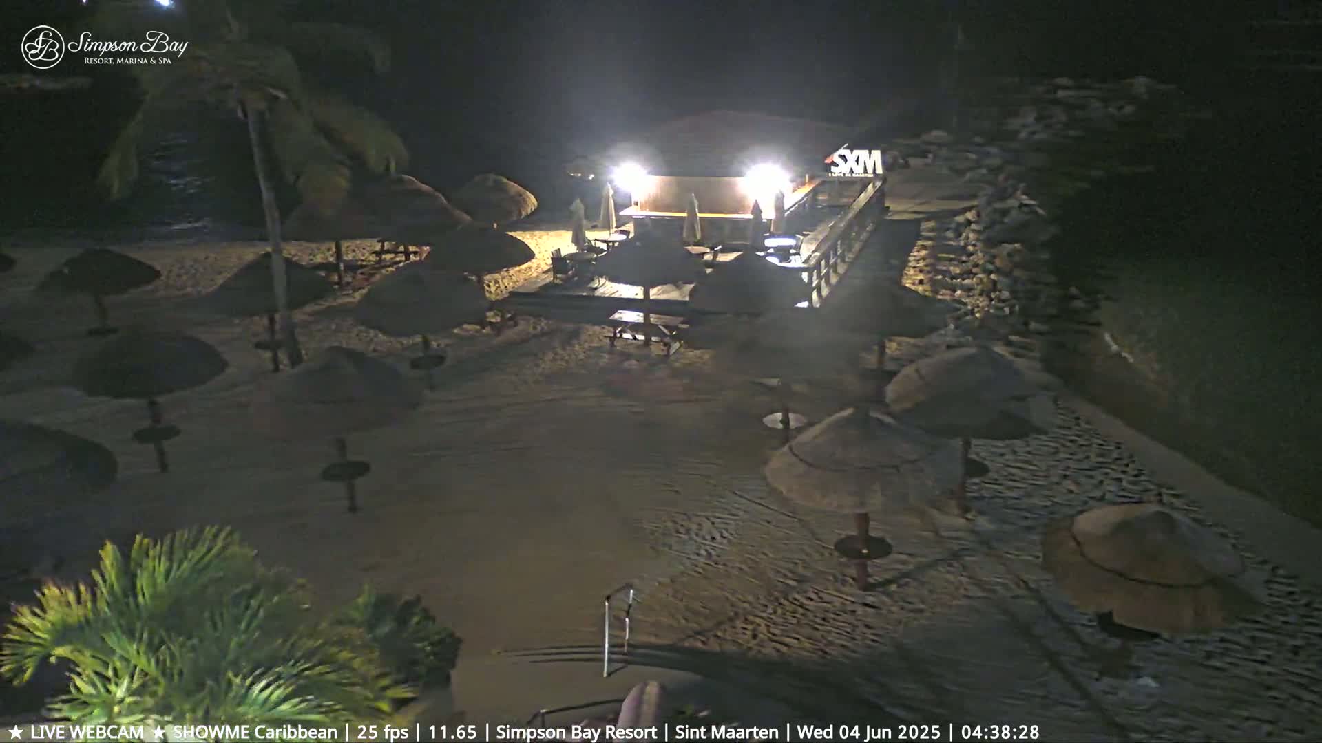 A nighttime beach scene shows a lighted beachfront bar and many closed thatched umbrellas on a sandy beach next to dark water.