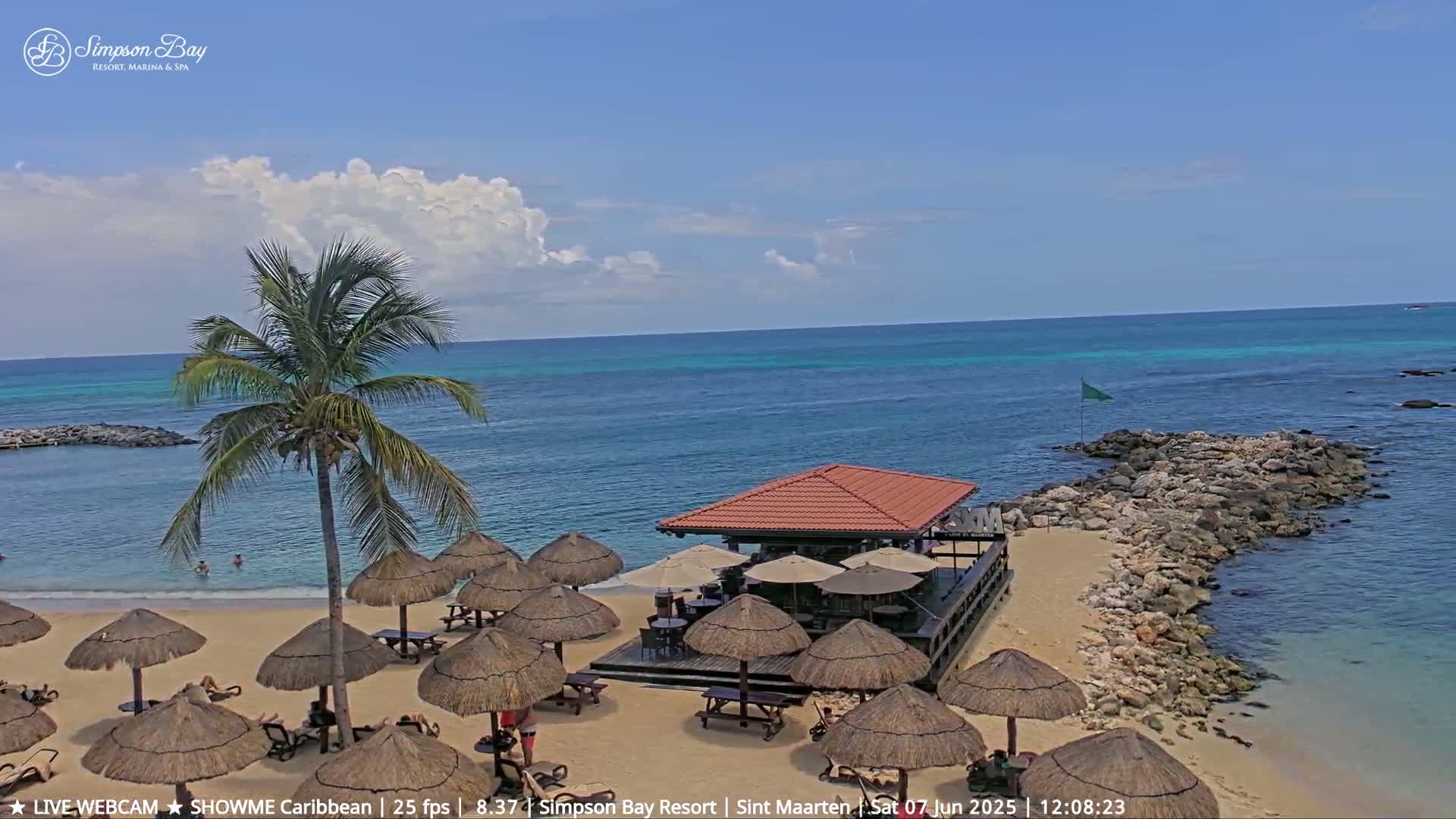 A beach bar with thatched umbrellas sits on a sandy beach next to a calm, turquoise ocean under a partly cloudy sky.