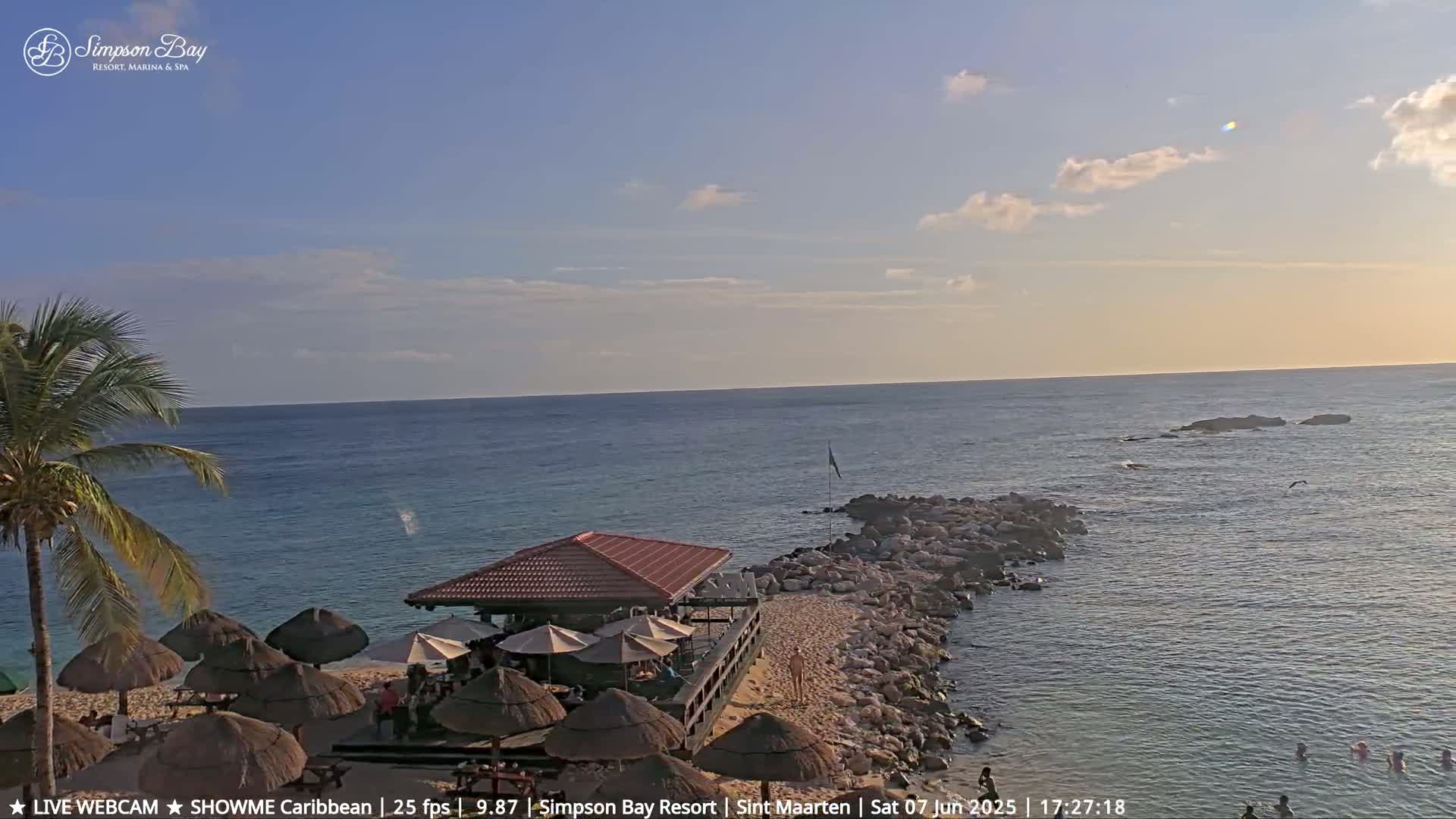 A partly cloudy sunset over a calm ocean shows a beach bar with thatched umbrellas and a rocky jetty with people swimming in the water.