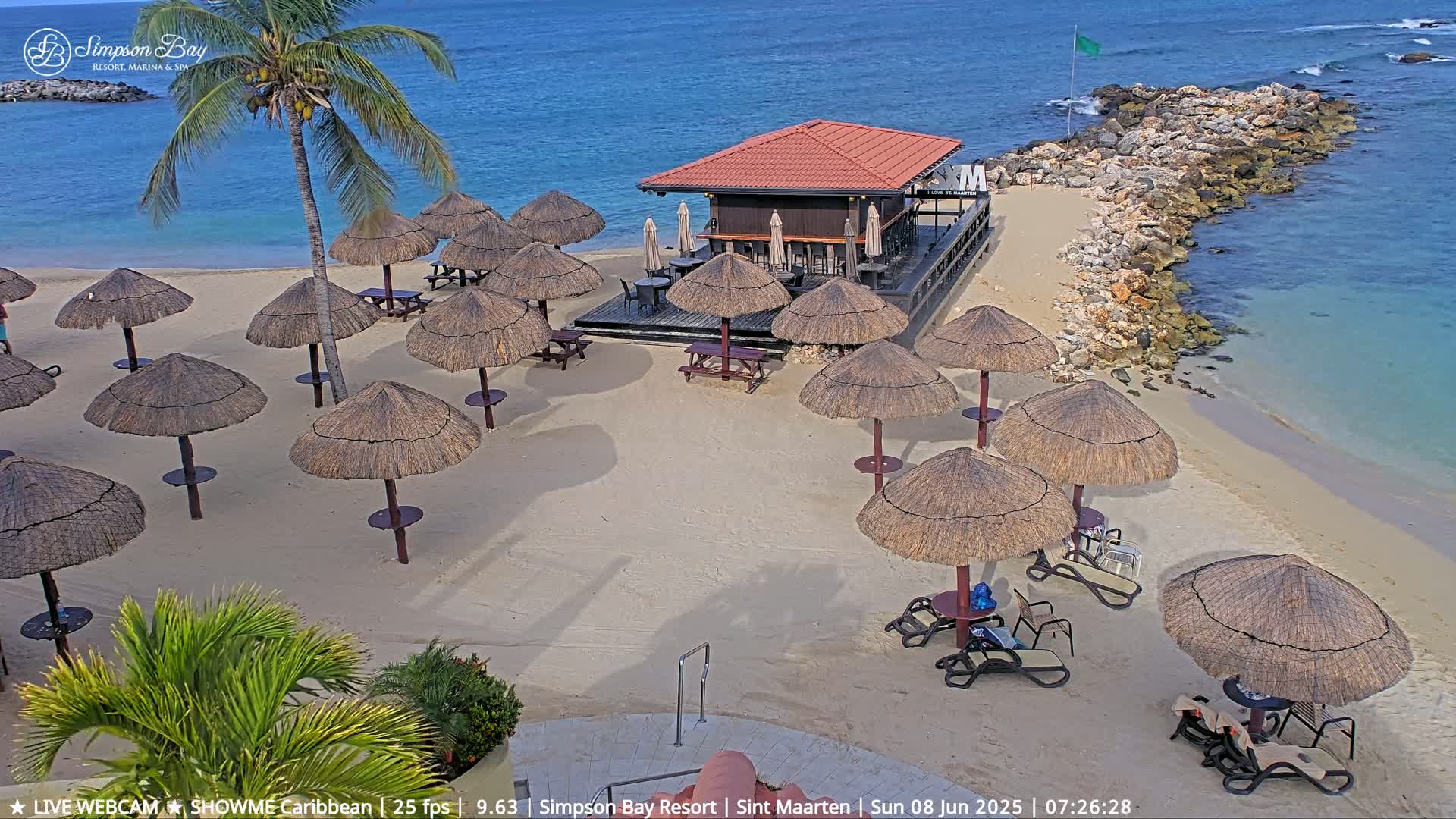 A sandy beach with numerous thatched umbrellas, beach chairs, and tables surrounding a small bar on a sunny day near calm, clear water.