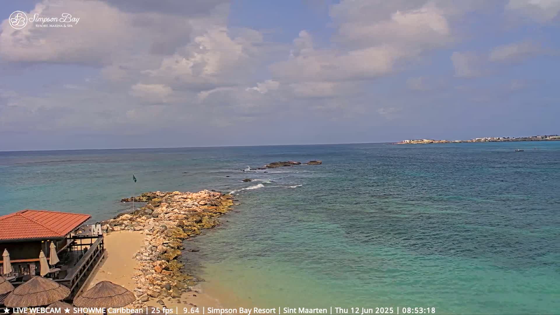 A partly cloudy day shows a rocky shoreline with a small building and beach area next to clear, turquoise water extending to a distant landmass.