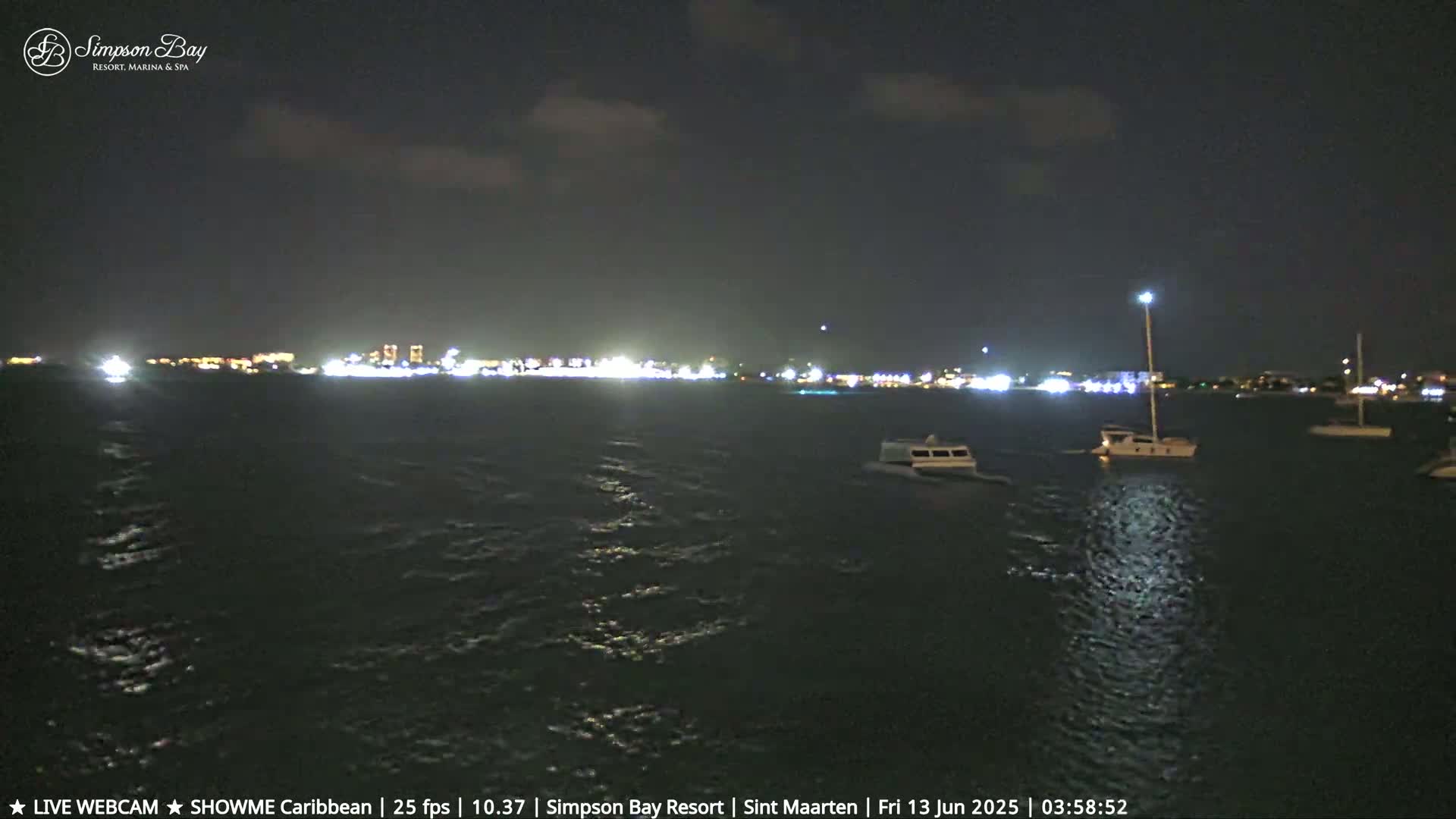 At night, several boats are moored in calm water with a brightly lit town visible in the distance.