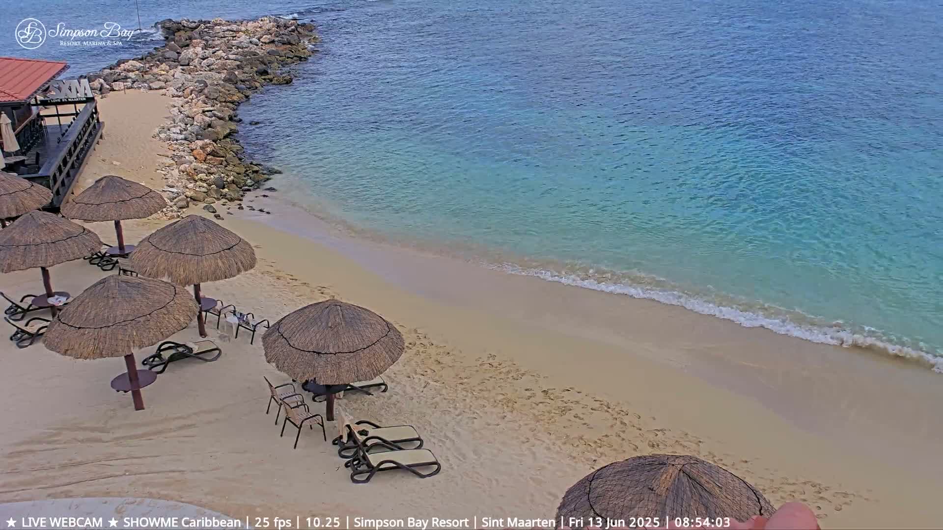 A sandy beach with calm, clear turquoise water and several thatched umbrellas and lounge chairs sits next to a rocky shoreline.