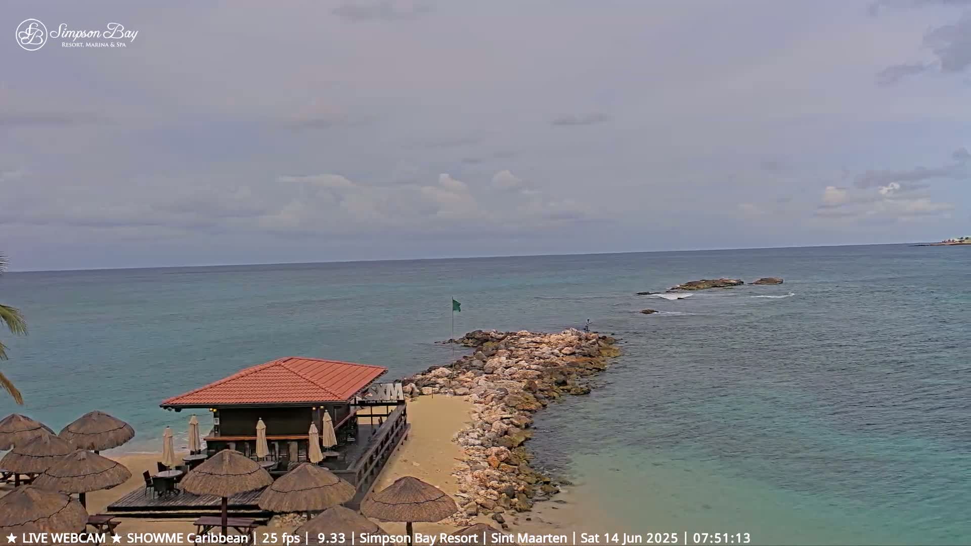 A beach bar with thatched-roof umbrellas sits on a sandy beach next to a rocky jetty extending into a calm, turquoise ocean under a partly cloudy sky.