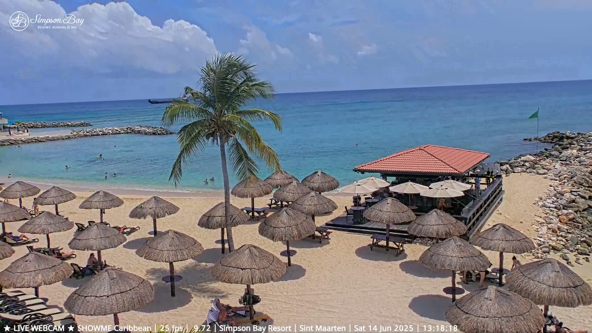 A sandy beach with numerous thatched umbrellas, a beachside bar, and calm turquoise water under a partly cloudy sky.
