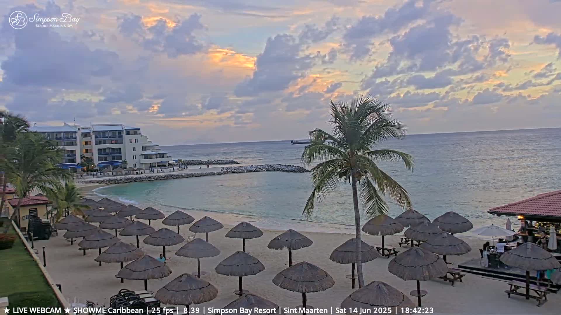 A sandy beach with numerous thatched umbrellas, a beachside bar, and calm turquoise water under a partly cloudy sky.